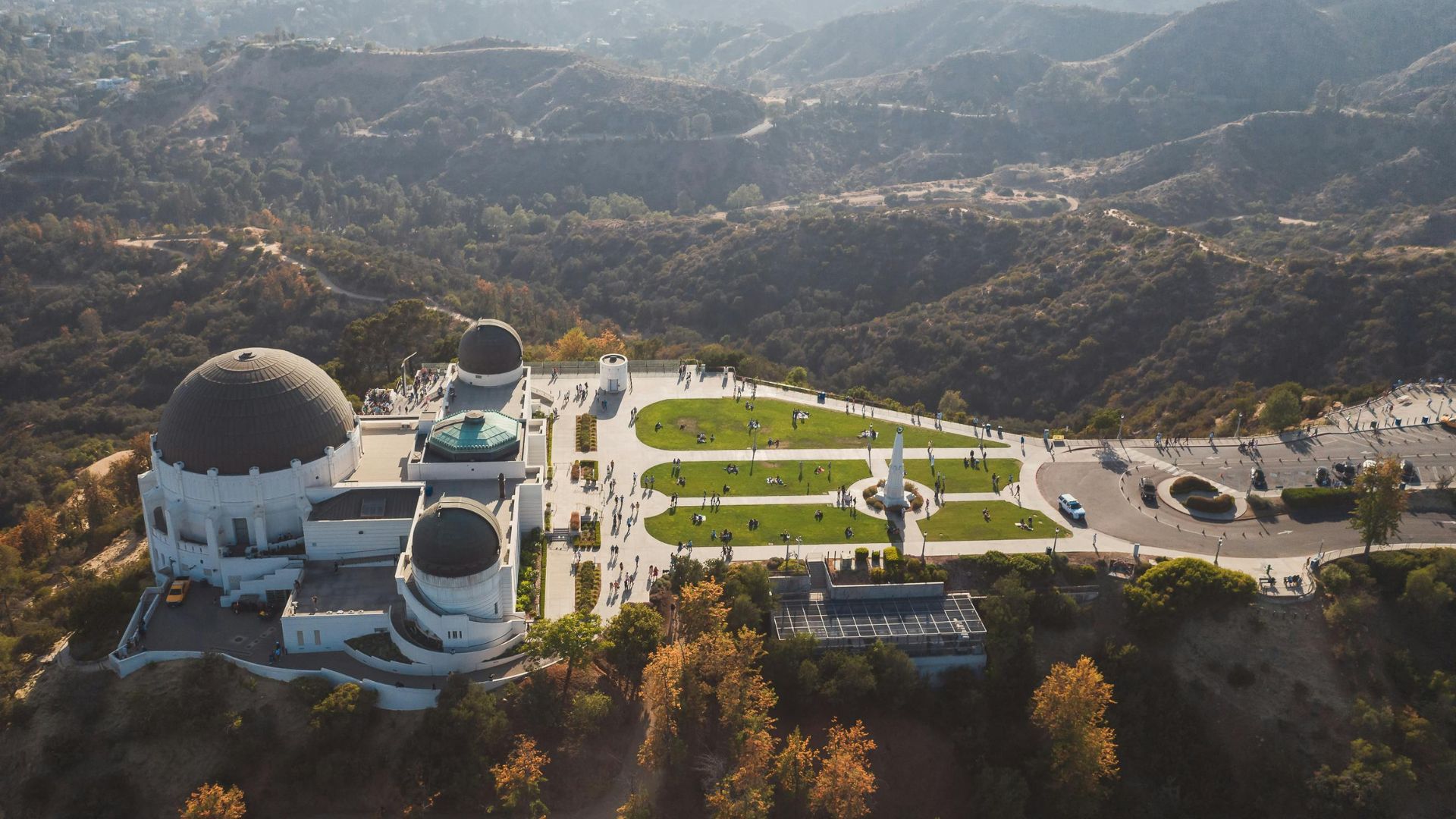 Aerial view of Griffith Observatory in Los Angeles, California, showcasing the iconic building, surrounding green lawns, and winding roads within Griffith Park, with the cityscape visible in the background. 