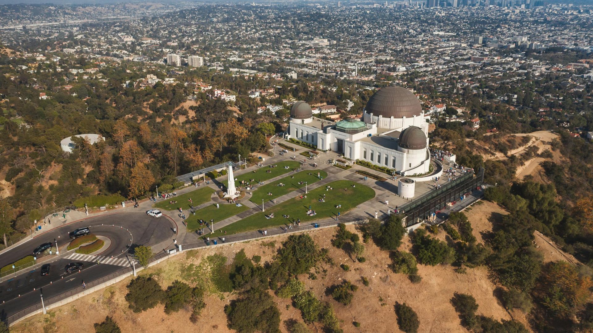 Aerial view of Griffith Observatory in Los Angeles, California, showcasing the iconic building, surrounding green lawns, and winding roads within Griffith Park, with the cityscape visible in the background. 