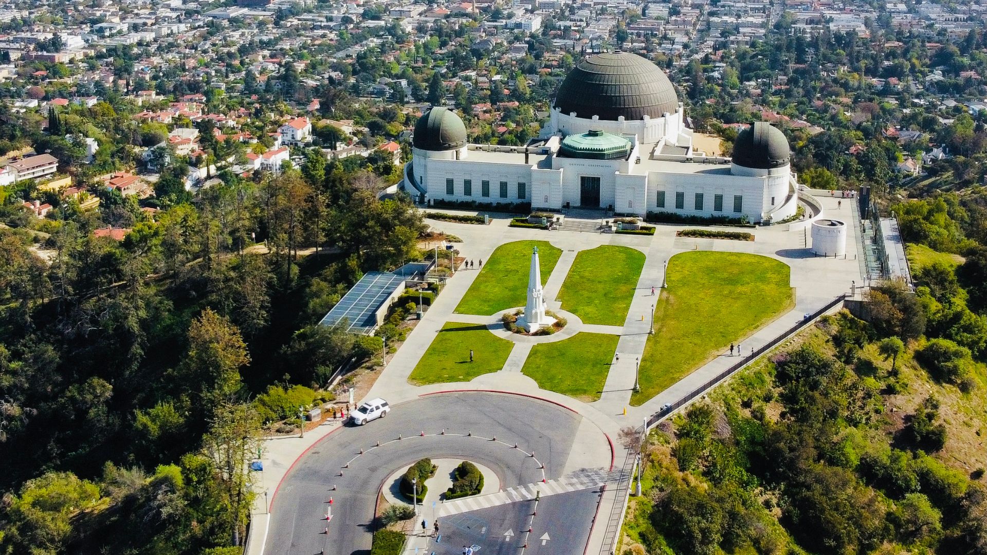 Aerial view of Griffith Observatory, a landmark in Los Angeles, California, showcasing its distinctive white building with domes, surrounding green spaces, and a portion of the city skyline in the background.