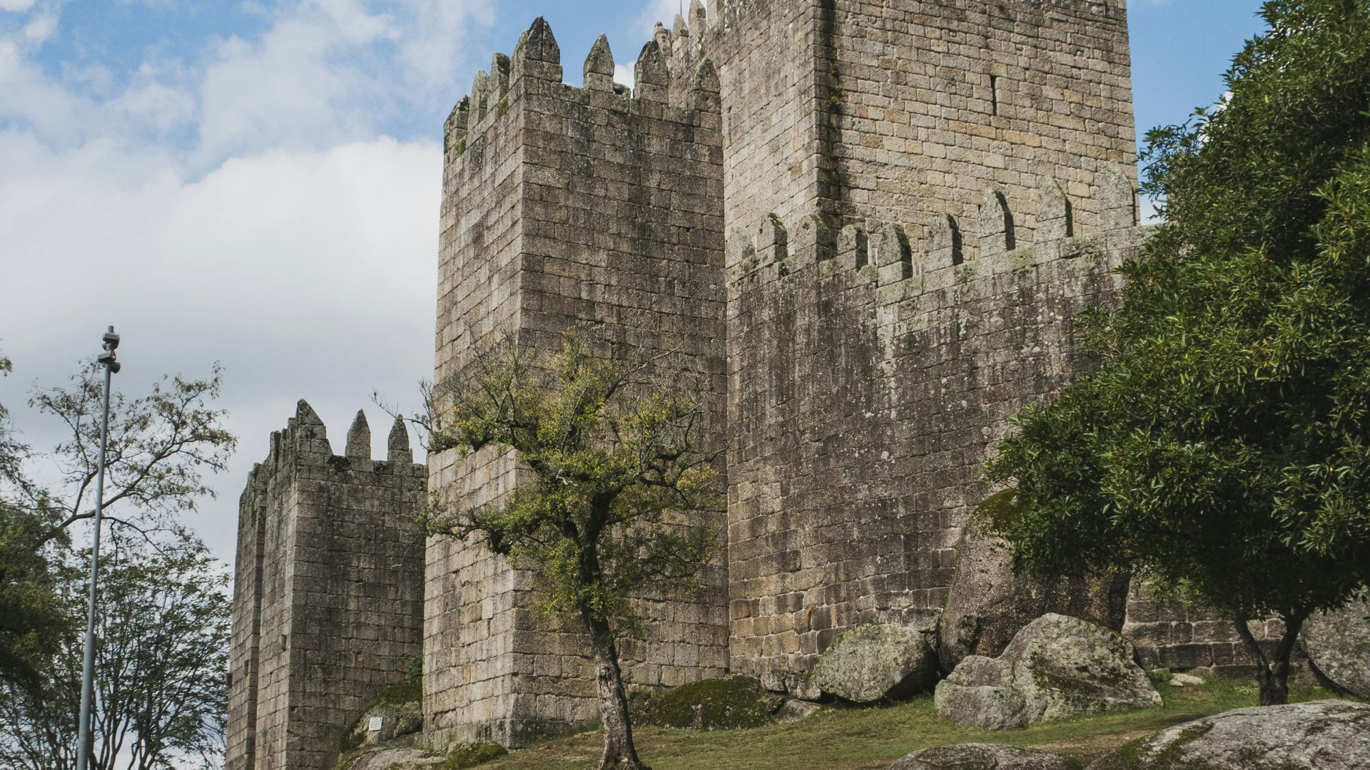 A large, historic stone castle with crenellated walls stands on a grassy hill under a blue sky with clouds.