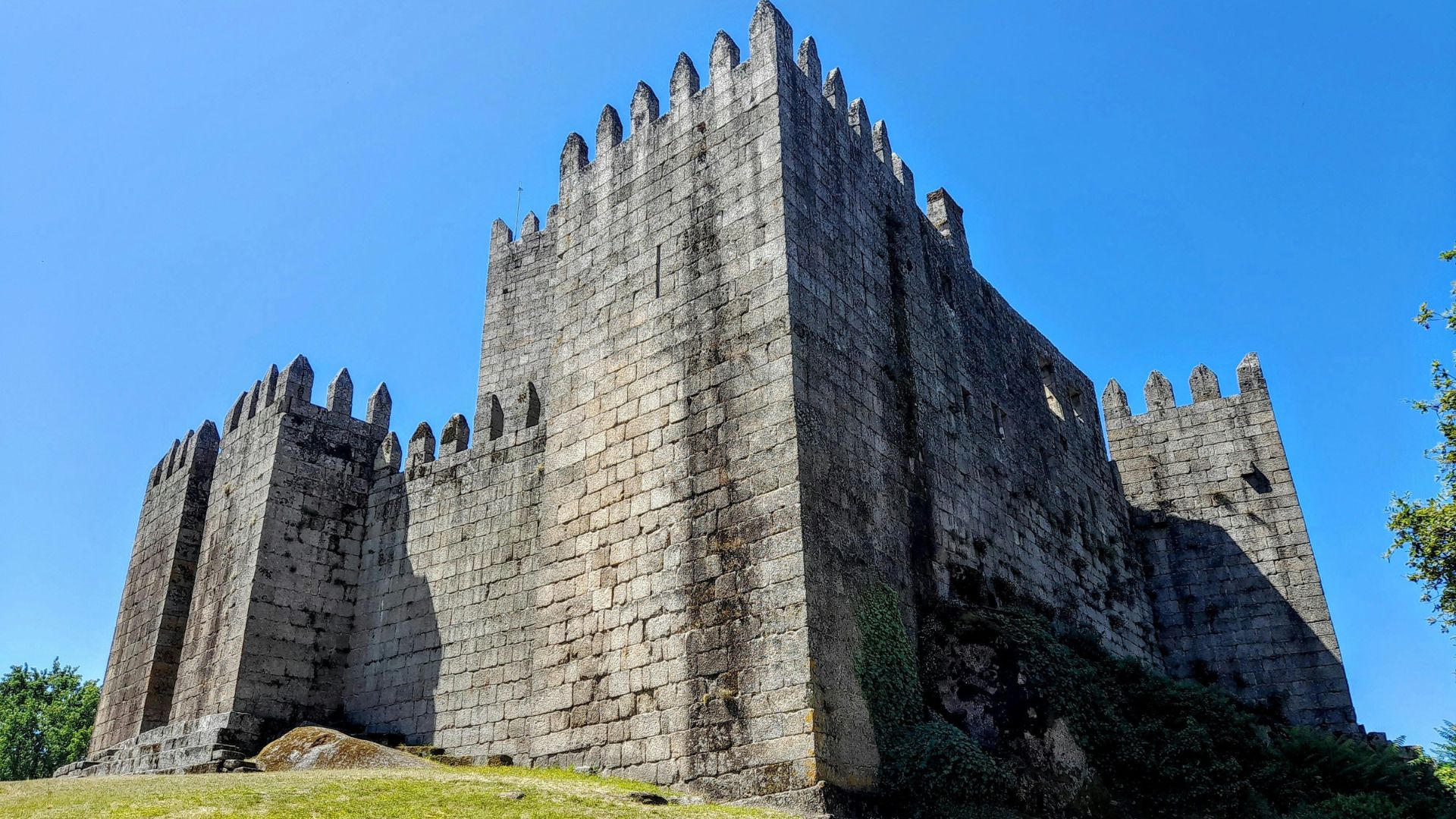 A low-angle shot of Guimarães Castle in Portugal, a medieval stone fortress with crenellated walls, situated on a grassy hill under an overcast sky.