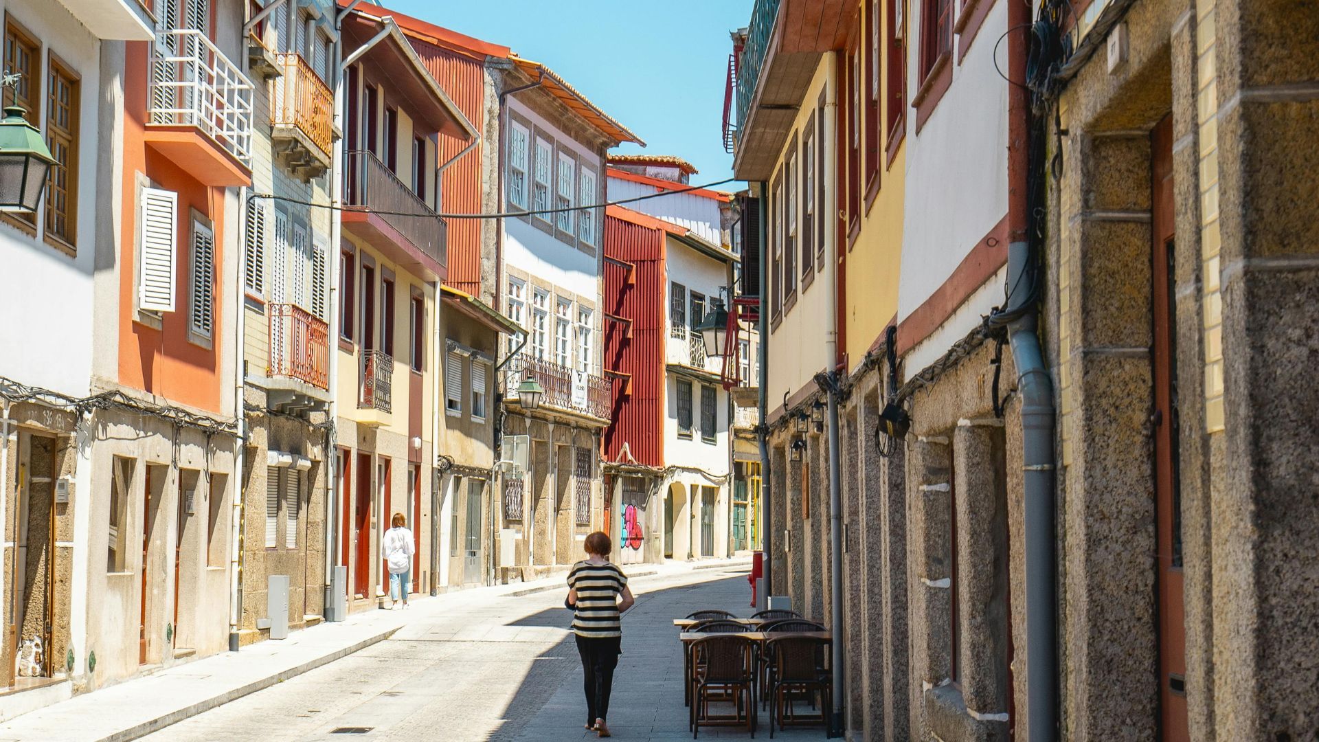 A narrow, historic street in Guimarães, Portugal, lined with traditional buildings featuring colorful facades, balconies, and stone foundations, with a person walking in the distance and tables set outside a building on the right.