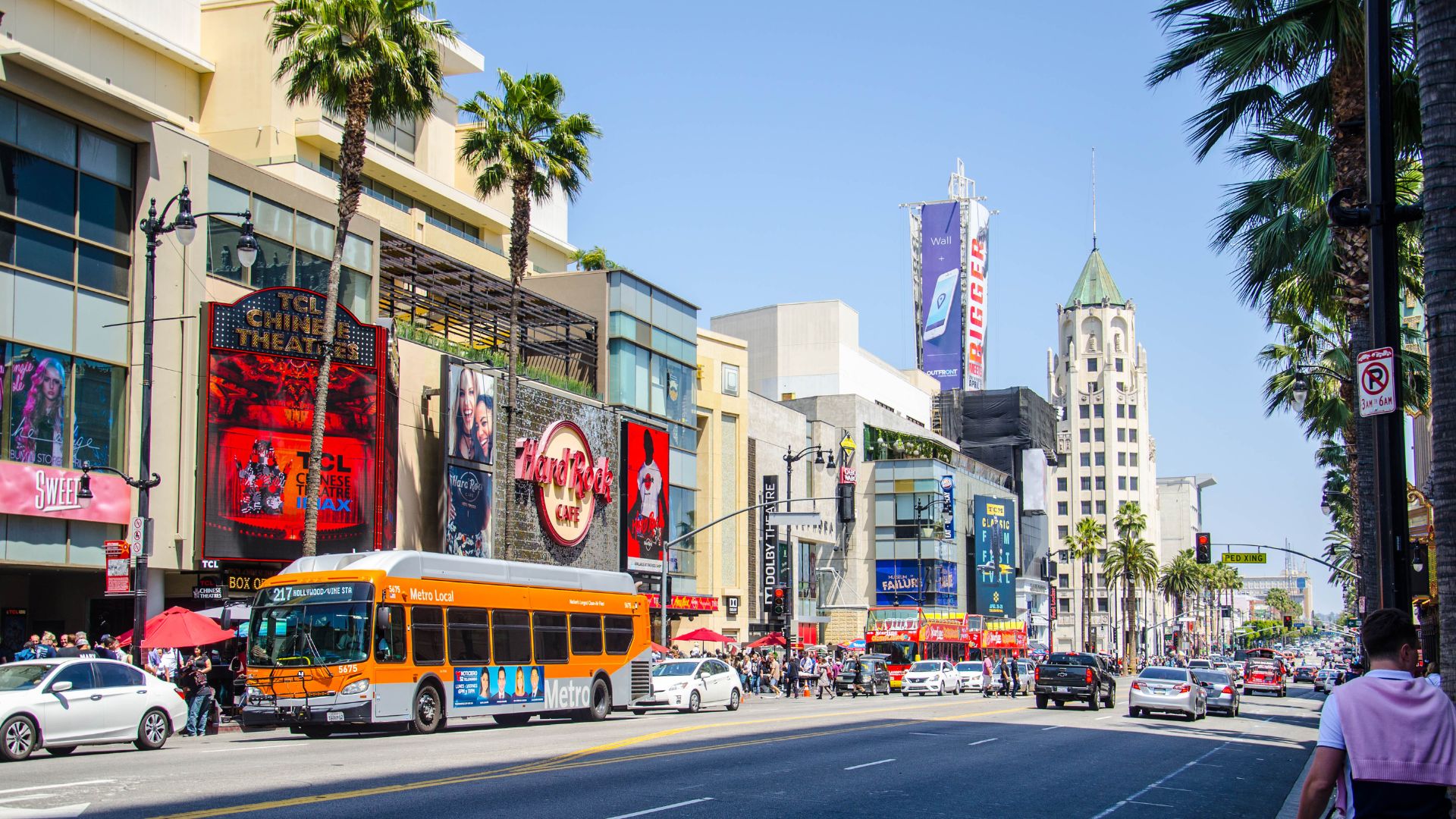 A vibrant daytime view of Hollywood Boulevard in Los Angeles, California, featuring iconic palm trees, the Hollywood sign, and bustling street activity.