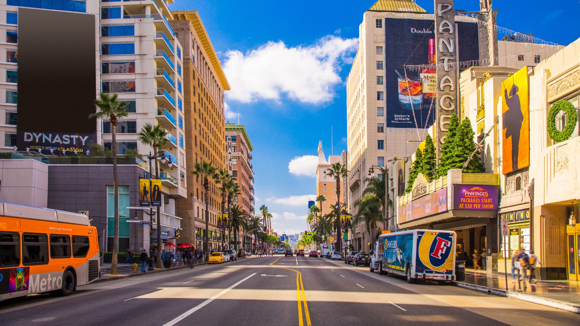 A vibrant daytime view of Hollywood Boulevard in Los Angeles, California, featuring iconic palm trees, the Hollywood sign, and bustling street activity.