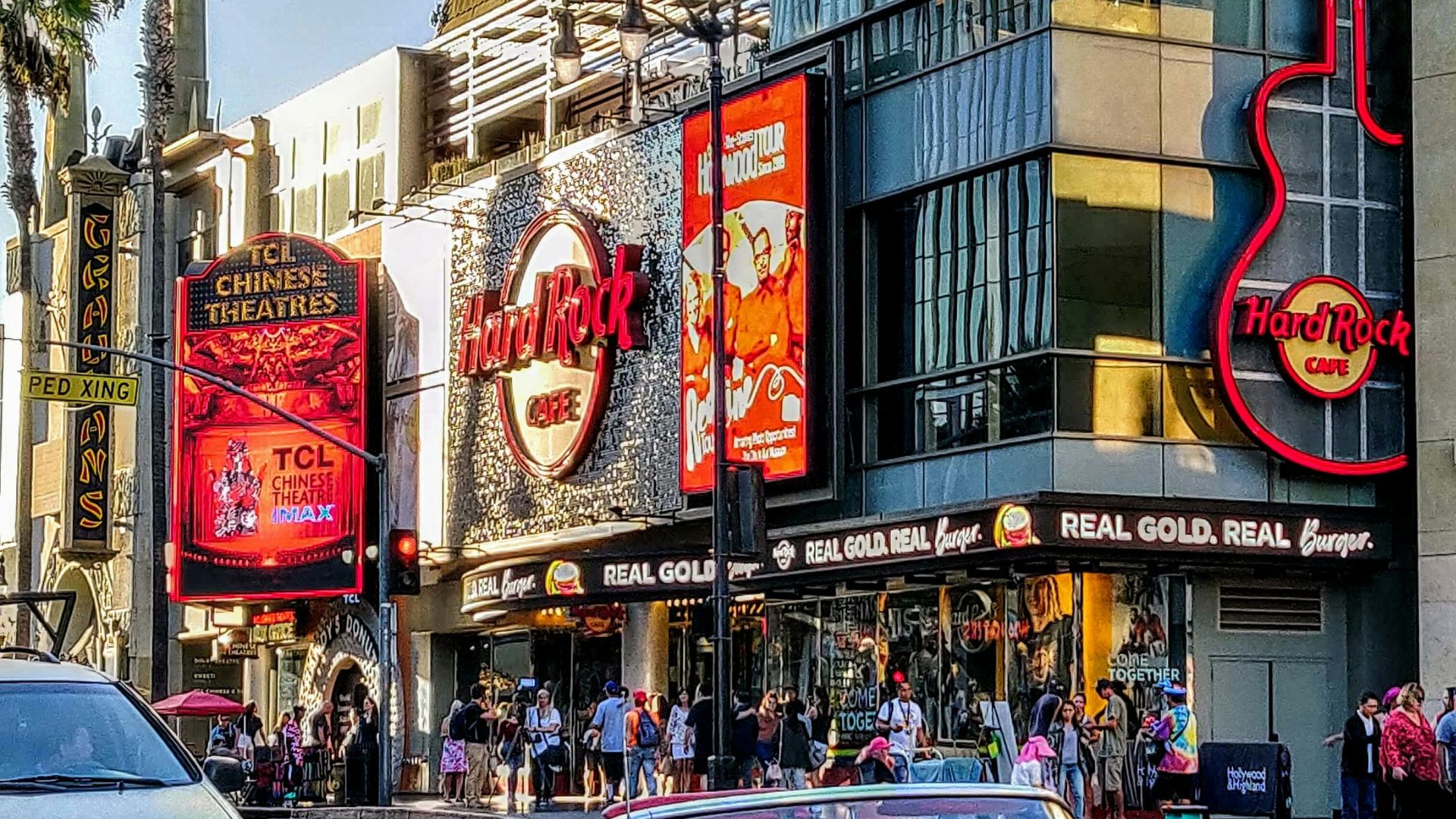 A vibrant street scene capturing the bustling energy of Hollywood Boulevard in Los Angeles, featuring the iconic Hard Rock Cafe with its large guitar sign and the TCL Chinese Theatre with its classic marquee.