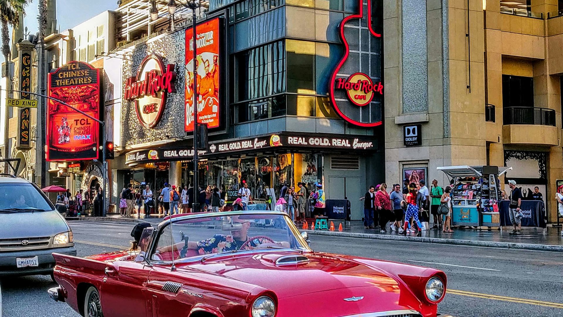 A vibrant street scene capturing the bustling energy of Hollywood Boulevard in Los Angeles, featuring the iconic Hard Rock Cafe with its large guitar sign and the TCL Chinese Theatre with its classic marquee.