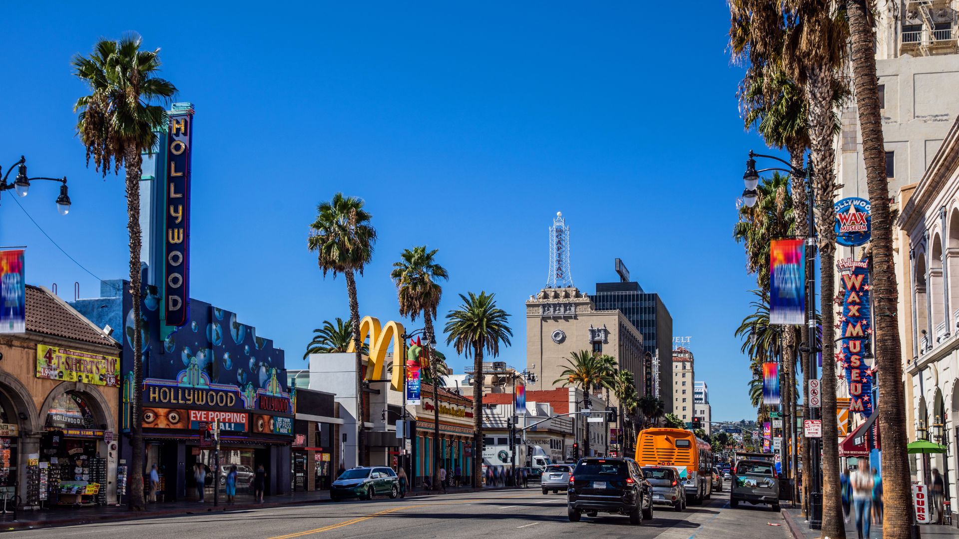 A vibrant daytime view of Hollywood Boulevard in Los Angeles, California, featuring iconic palm trees, the Hollywood sign, and bustling street activity.
