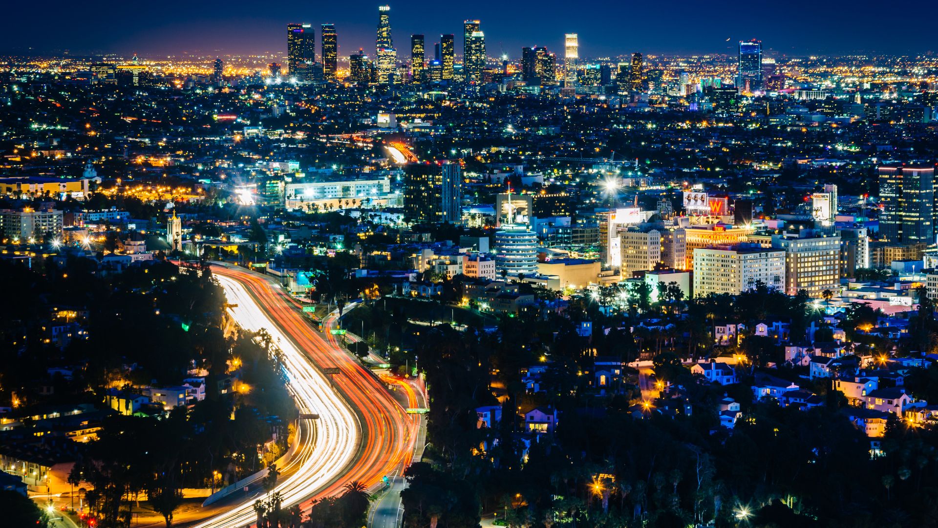 Nighttime view of Los Angeles skyline from the Hollywood Bowl Overlook. City lights illuminate the urban landscape, with a winding highway visible in the foreground. 