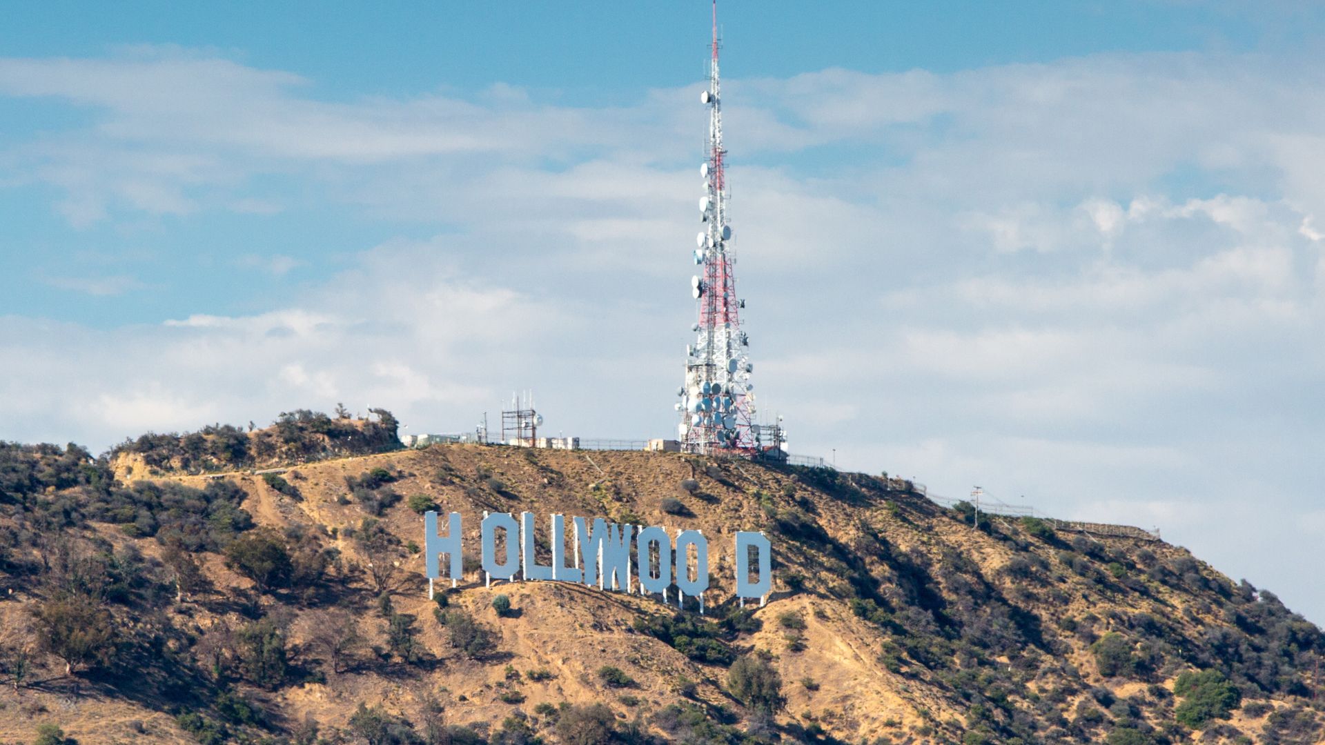 The iconic Hollywood Sign on Mount Lee, Los Angeles, with a radio tower behind it. Sparse vegetation and distant mountains are visible.