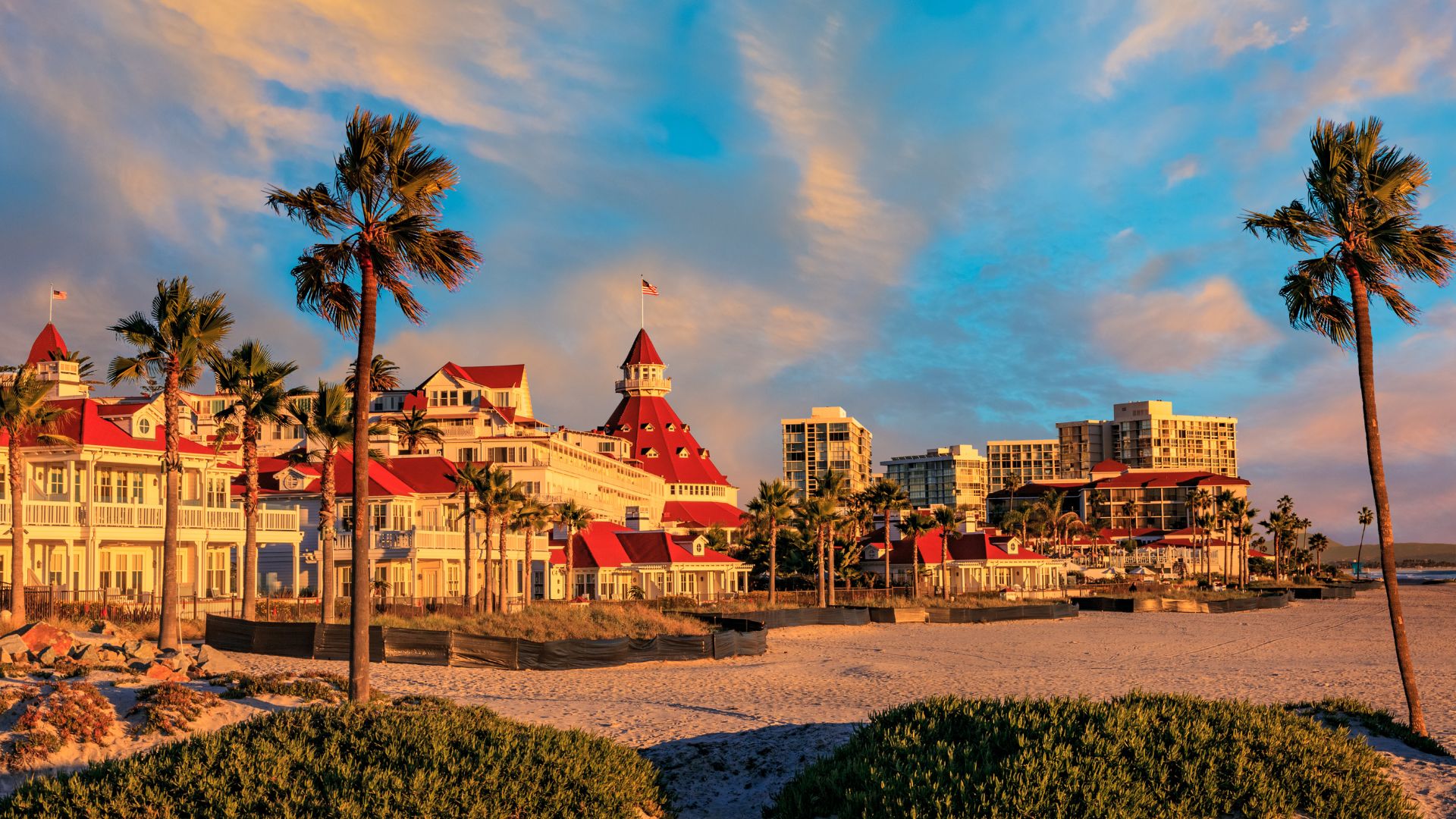 A historic, red-roofed Victorian hotel with palm trees on a sandy beach at sunset, with modern buildings in the background.