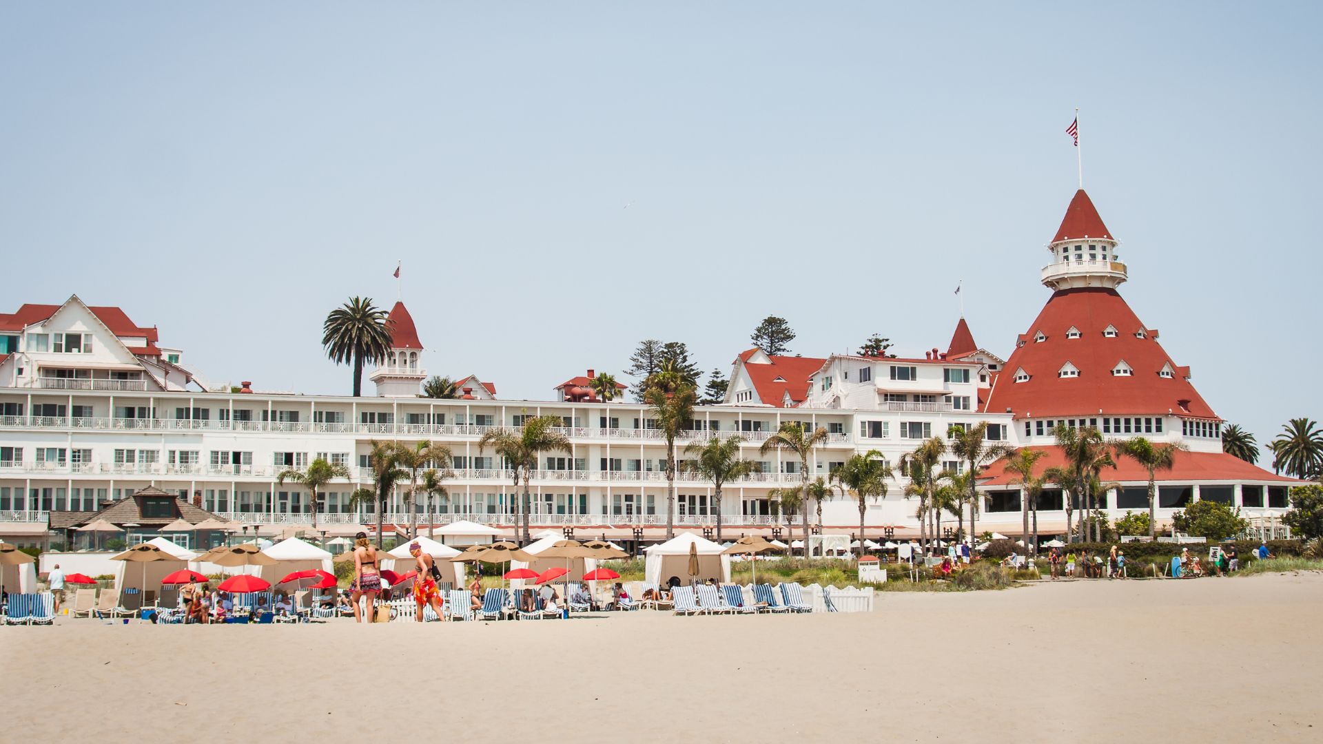 A wide shot of the iconic Hotel del Coronado, a large white Victorian-style hotel with distinctive red roofs and turrets, standing prominently on a sandy beach with numerous beach umbrellas and chairs, under a clear sky. Palm trees are visible around the hotel and on the beach.