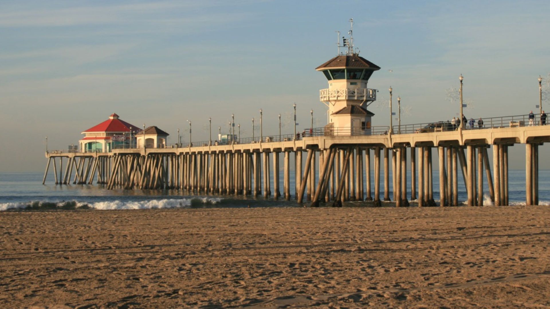 Huntington Beach Pier and iconic red-roofed building overlooking the Pacific Ocean with sandy beach and distant city skyline in Huntington Beach, California, a popular surf destination.