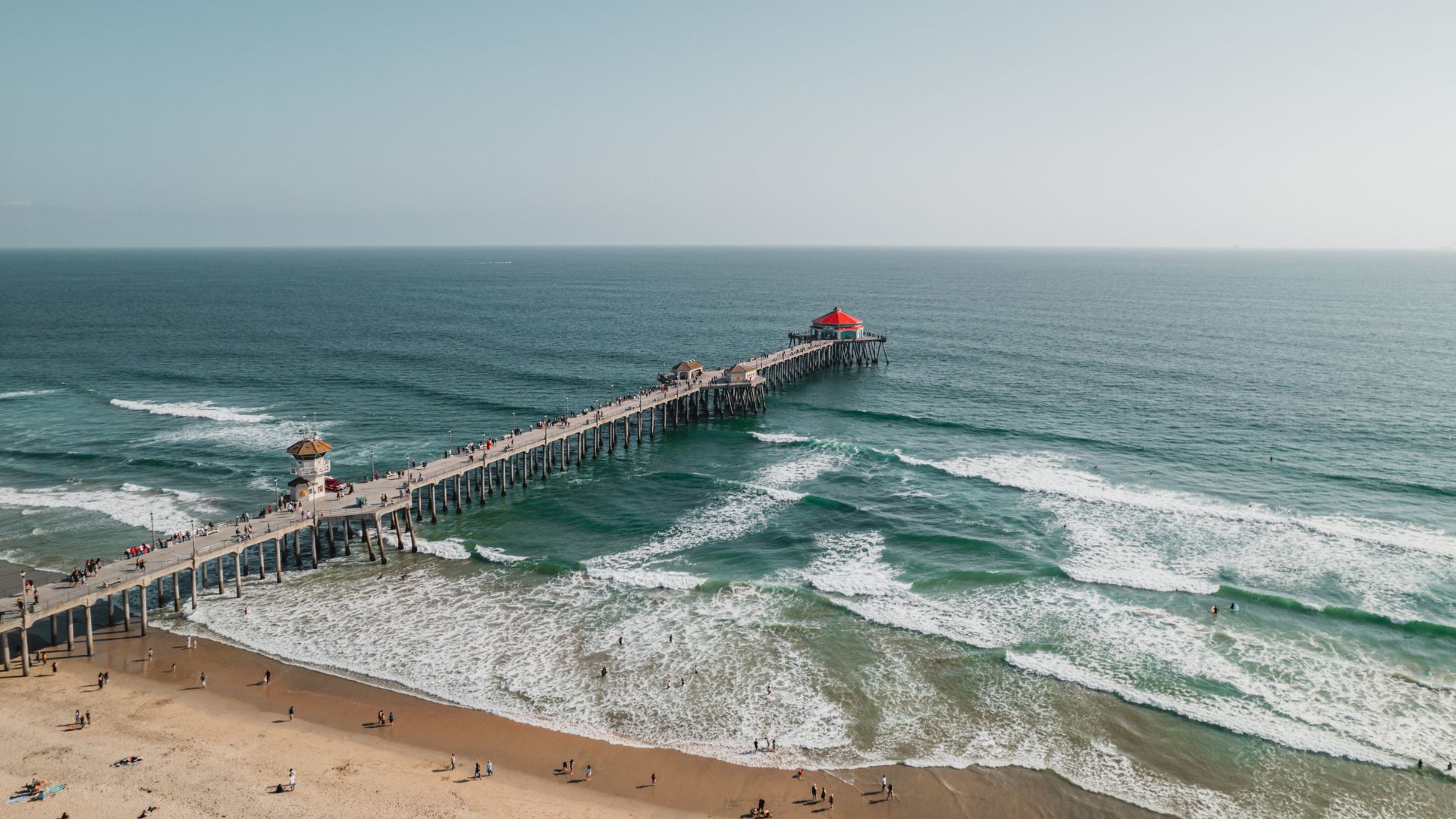 A long pier, the Huntington Beach Pier, extends into the Pacific Ocean under a partly cloudy sky. The pier features a prominent hexagonal building with a red roof and white walls near its end.