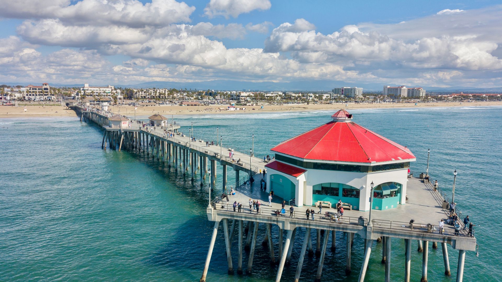 A long pier, the Huntington Beach Pier, extends into the Pacific Ocean under a partly cloudy sky. The pier features a prominent hexagonal building with a red roof and white walls near its end.