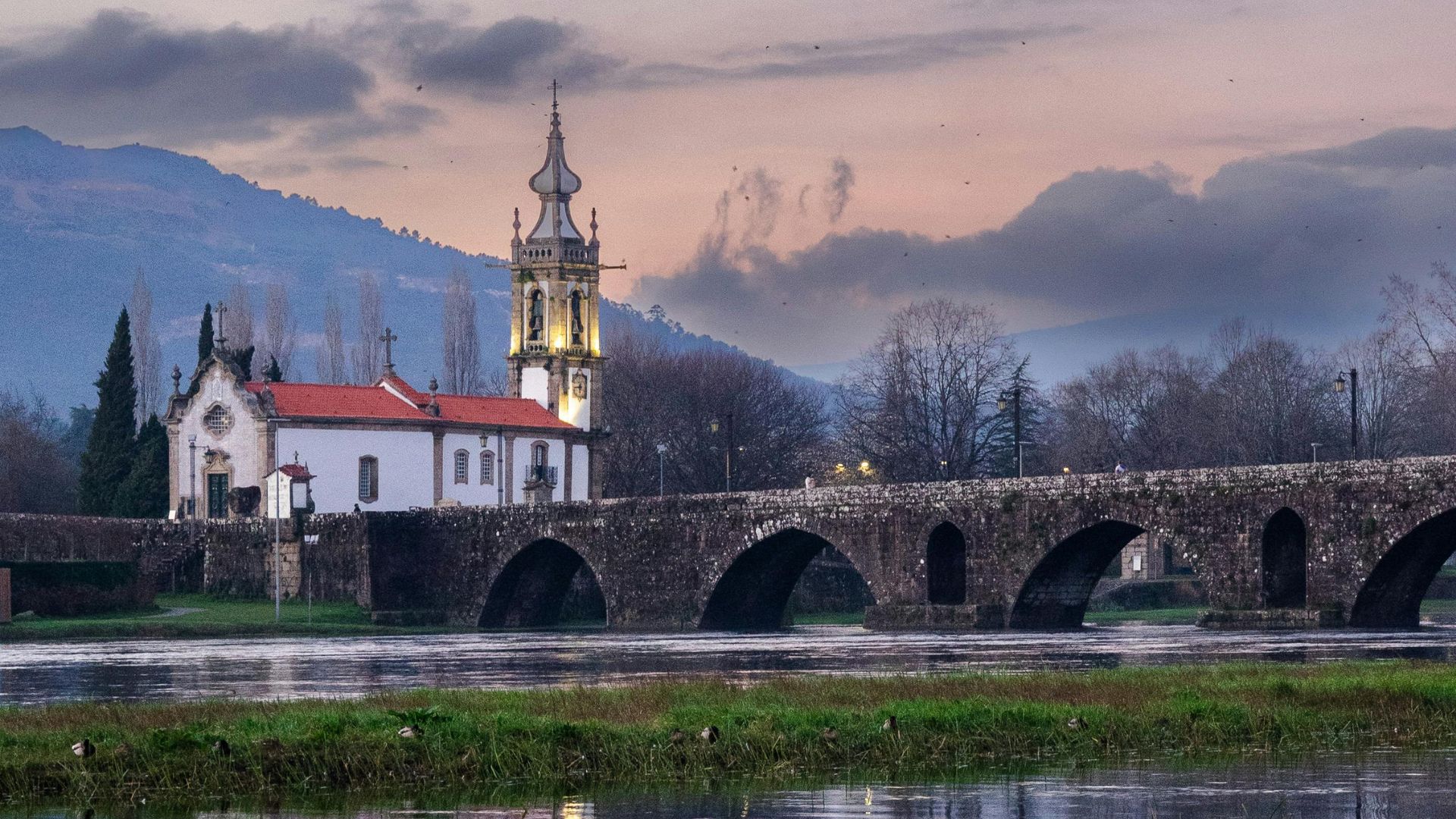 A picturesque view of the medieval bridge and the Igreja Matriz de Ponte de Lima reflected in the calm waters of the Lima River at dusk, with mountains in the background.
