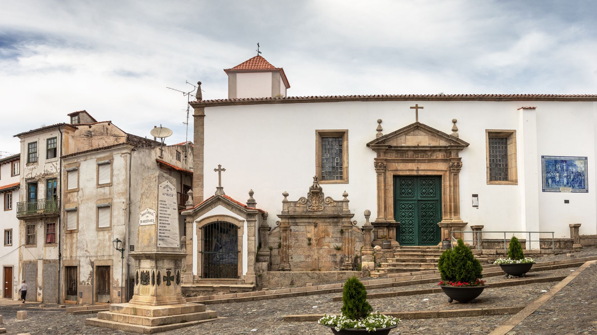A white church with a prominent stone entrance and green doors, featuring a small chapel to its left and a stone fountain in front, situated on a cobblestone street with planted shrubs in pots.