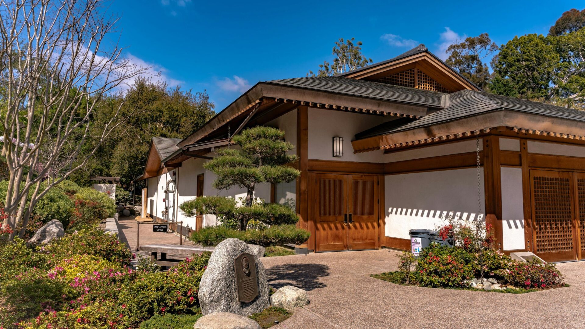 A traditional Japanese-style building with a wooden facade and tiled roof, situated within a landscaped garden featuring manicured trees, rocks, and flowering bushes under a clear blue sky.