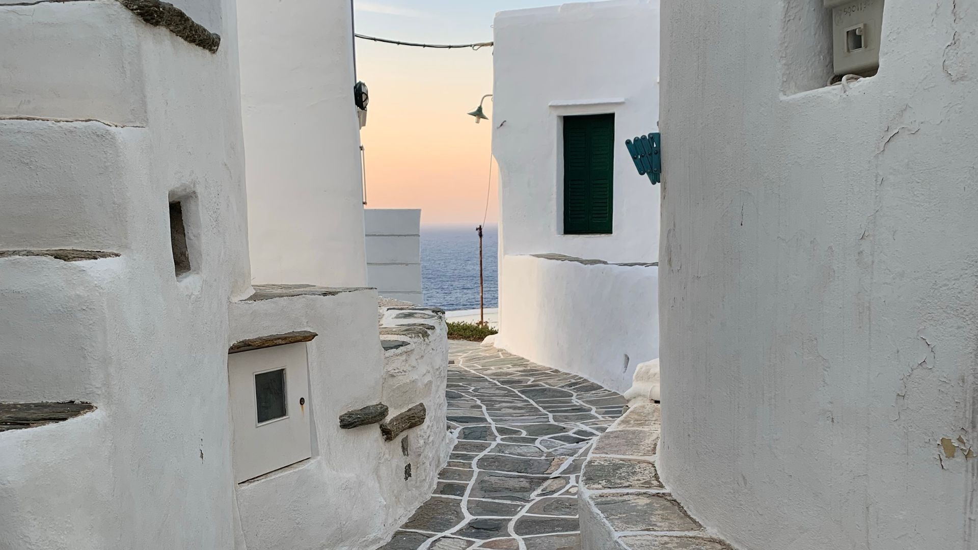 A narrow cobblestone alleyway lined with white buildings in Sifnos, Greece, leading to a sea view.