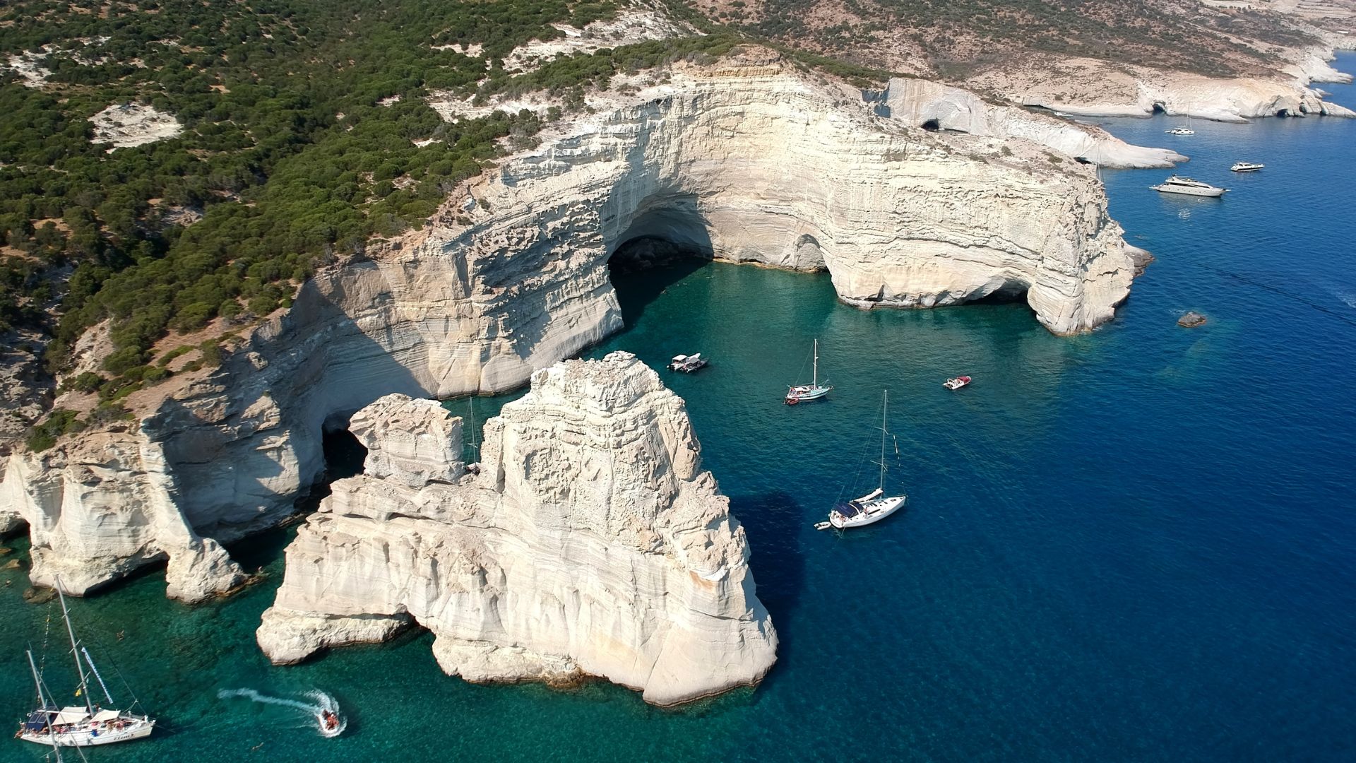 Aerial view of Kleftiko Beach in Milos, Greece, featuring white cliffs, caves, and clear blue waters with boats.
