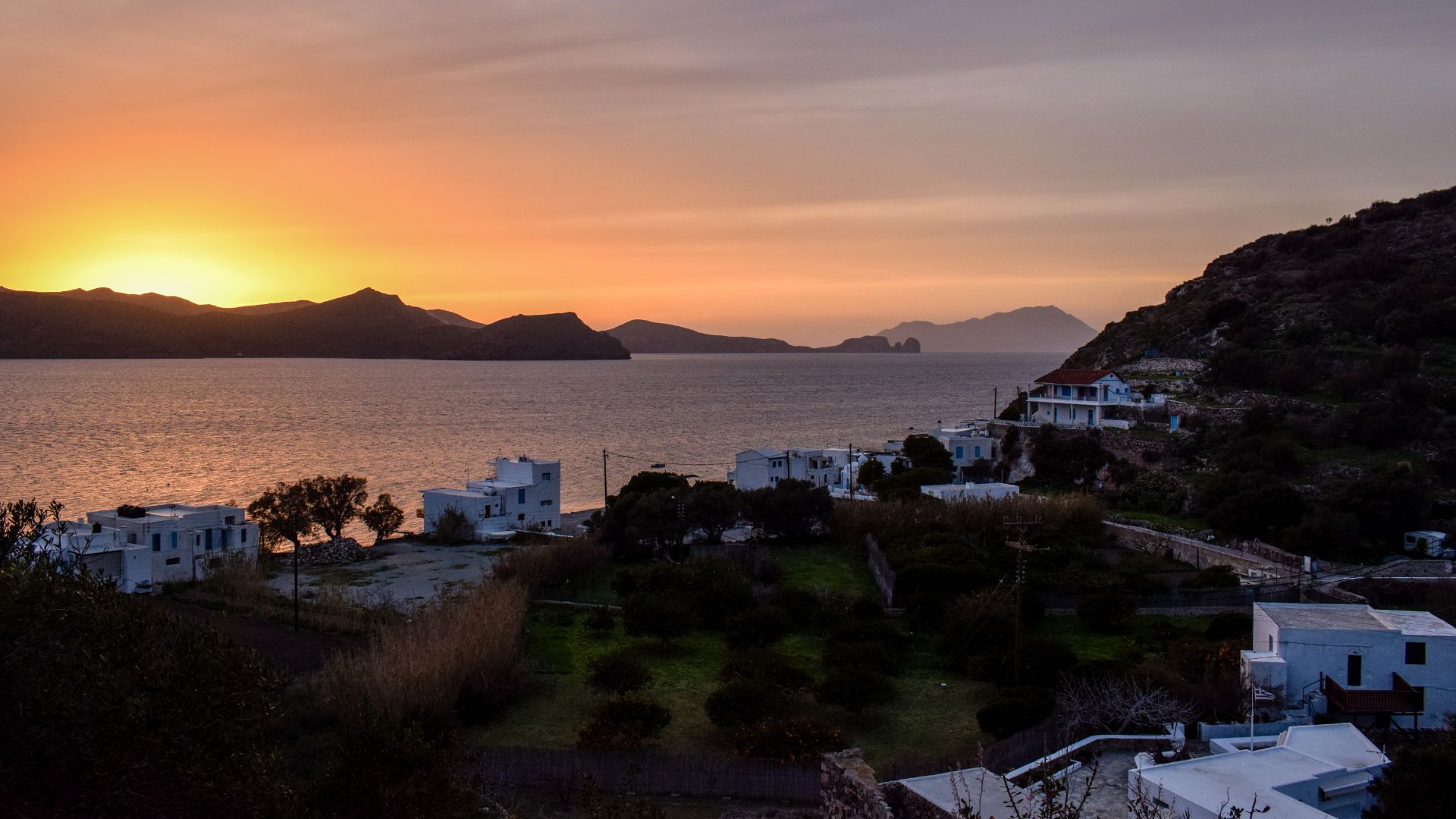 Sunset view of Klima Village in Milos, Greece featuring white buildings and greenery fscing the sea.