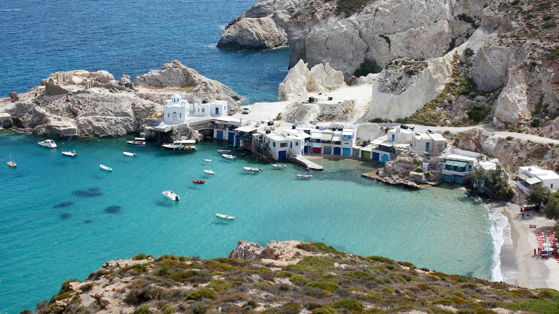 Aerial view of Klima village of Milos Island in Cyclades, Greece featuring white buildings with colorful doors, white rocky cliffs, and turquoise water with boats.