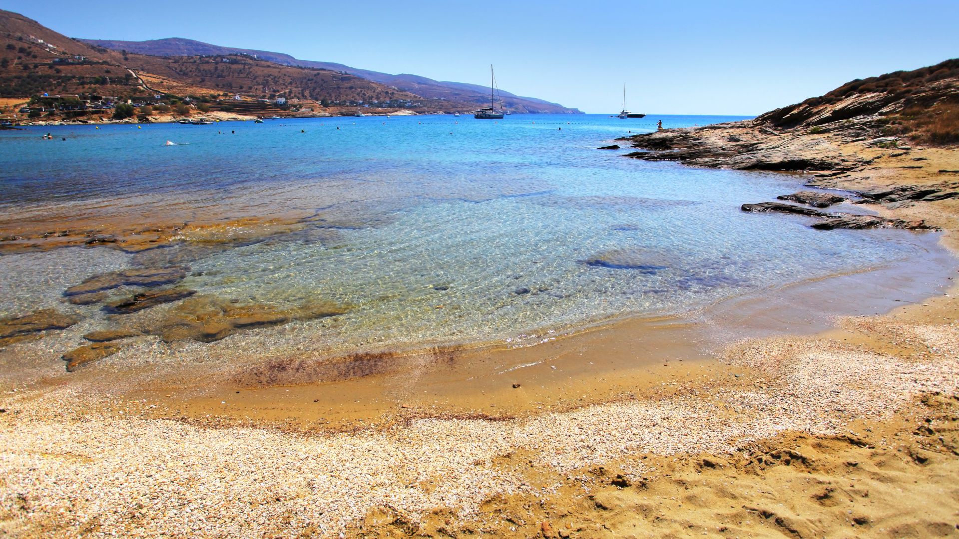 A picturesque view of Koundouros Beach in Kea, Greece, featuring clear turquoise water, sandy shore, and distant boats.