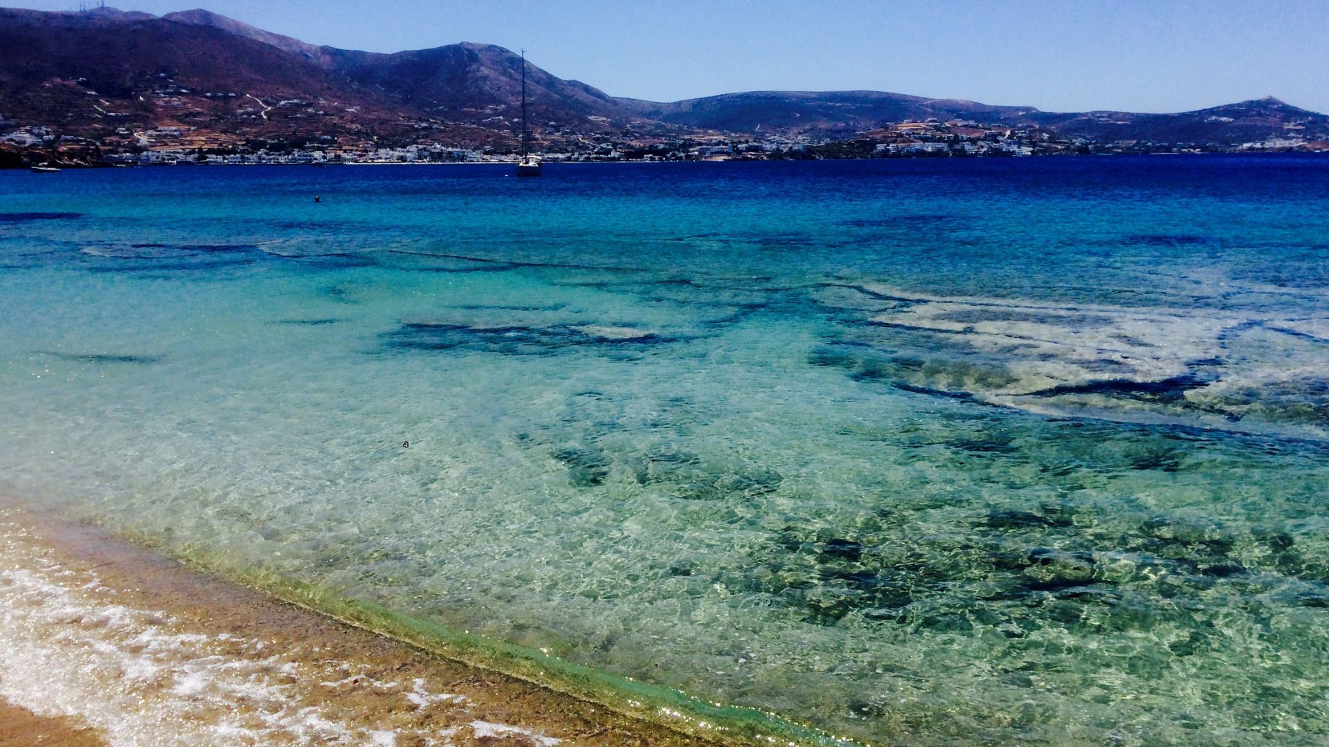Clear turquoise waters meet a sandy shore at Krios Beach, Paros, with mountains in the background.
