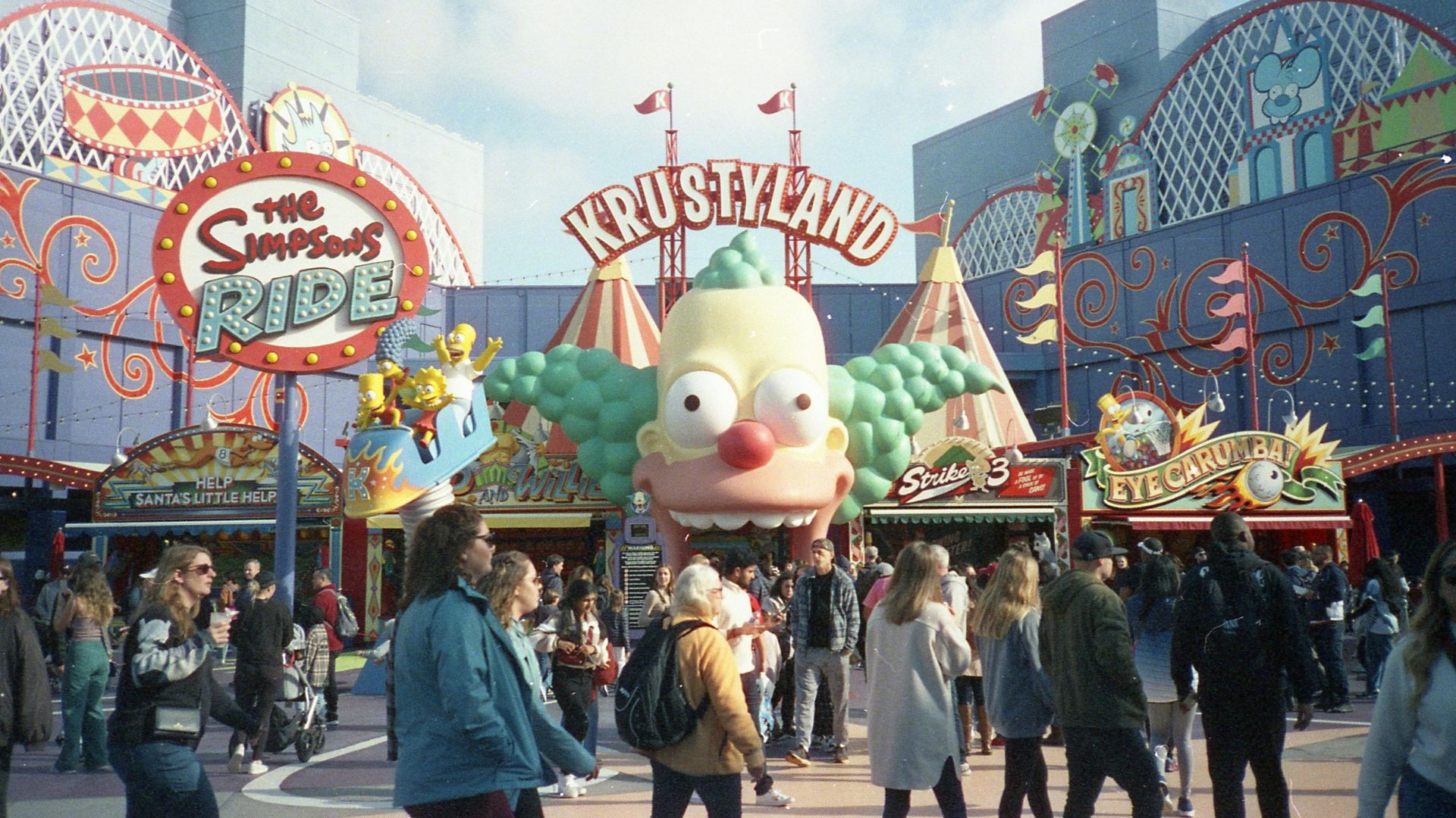 The entrance to Krustyland at Universal Studios Hollywood, featuring the iconic Krusty the Clown face, with guests entering the park. The Simpsons Ride is a popular attraction.