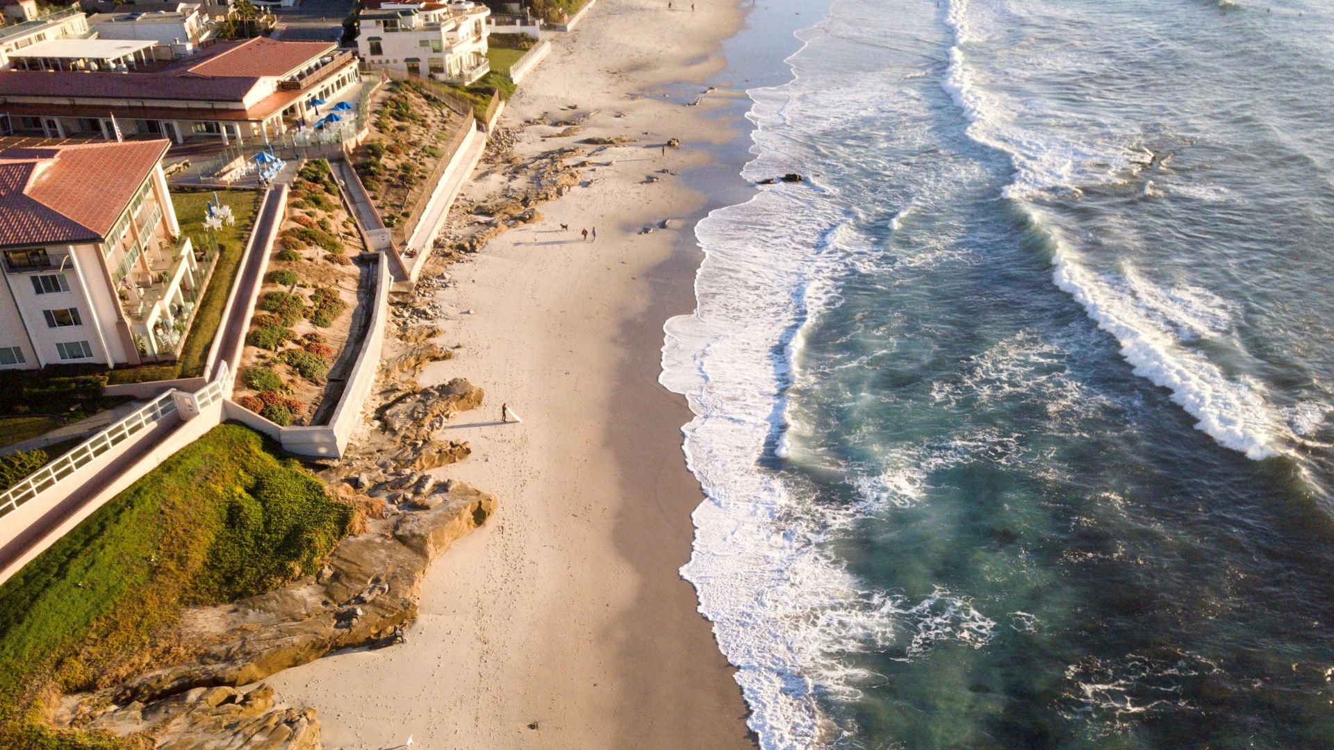 Aerial view of a sandy beach with crashing waves and coastal cliffs, featuring buildings and lush green areas along the shoreline.