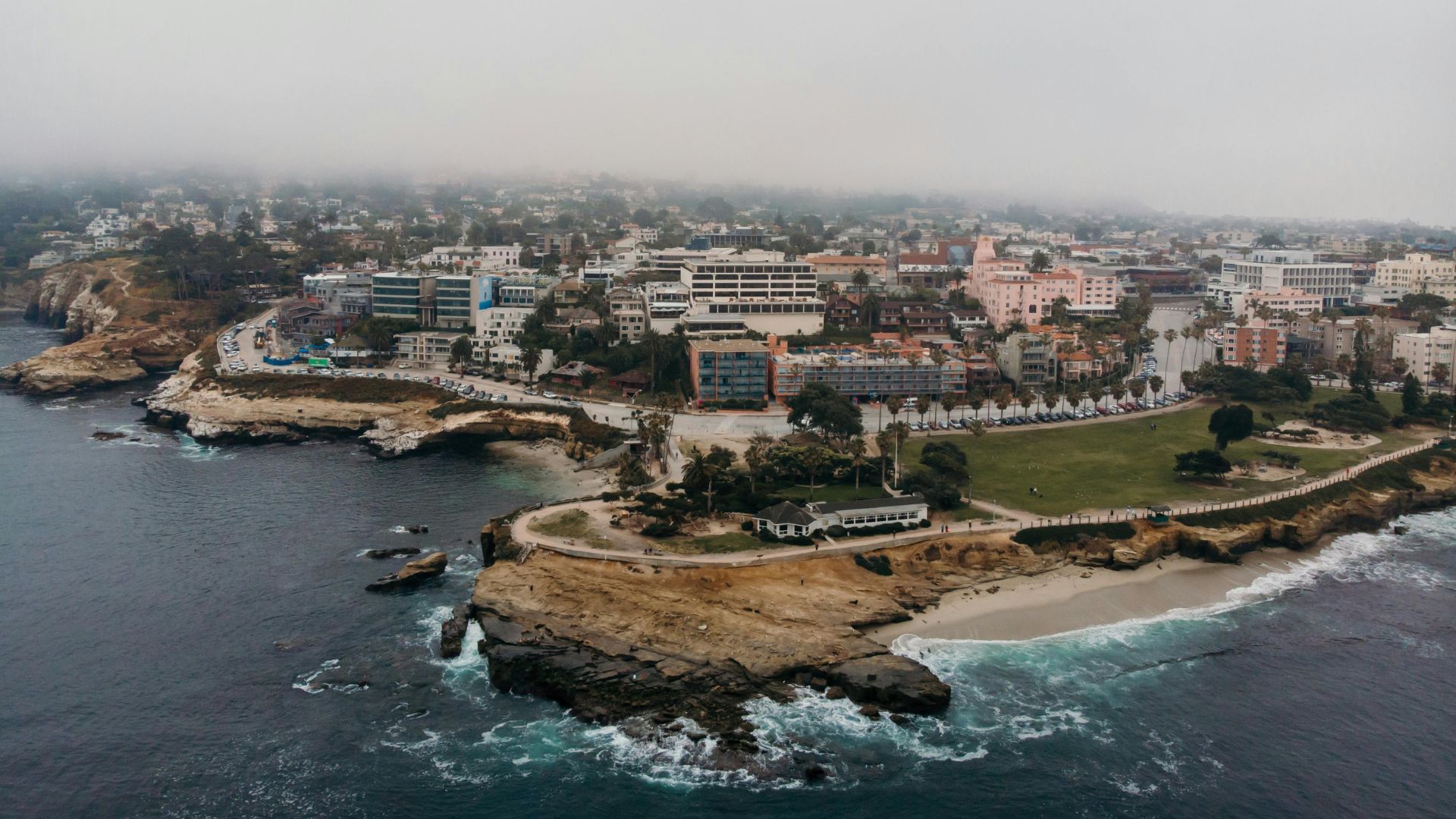 Aerial view of La Jolla Cove in San Diego, California, featuring coastal cliffs, a sandy beach, and buildings along the shoreline under a cloudy sky. 
