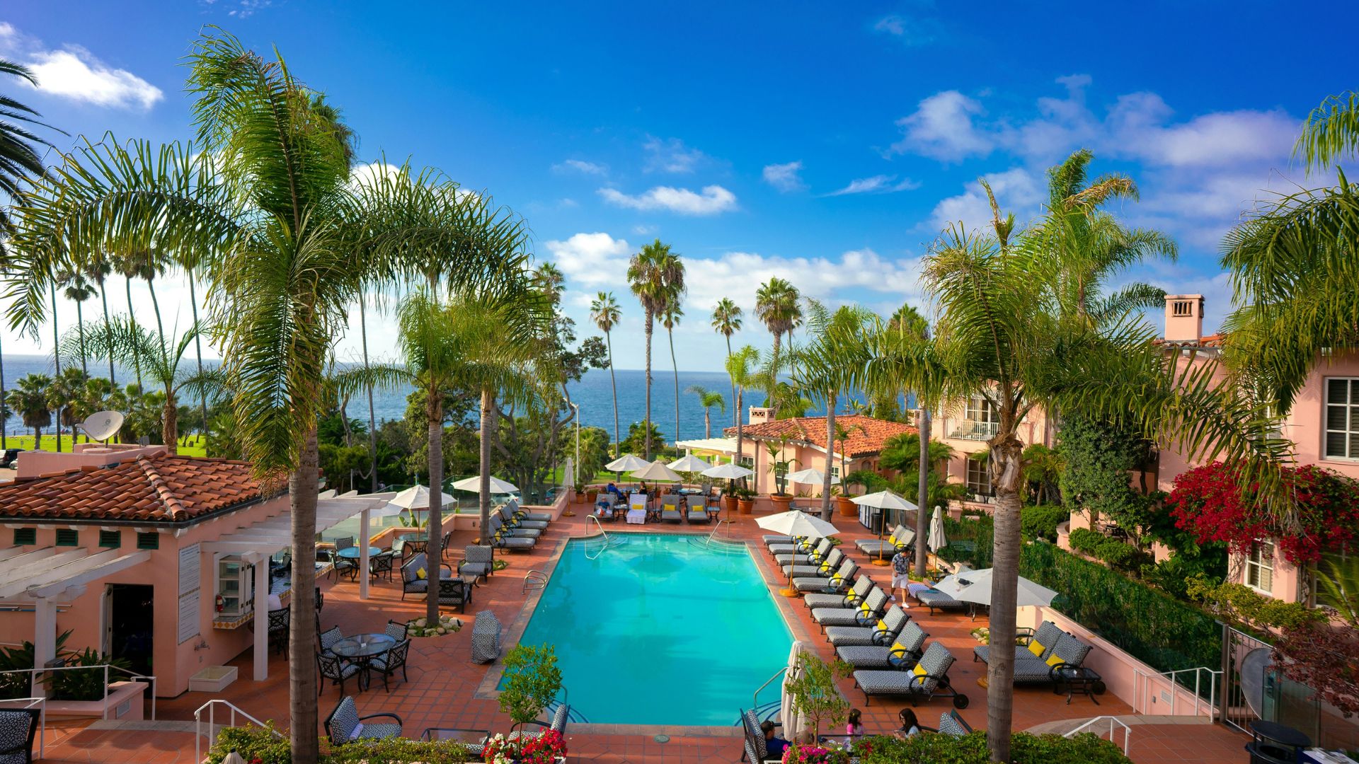 An aerial view of the La Valencia Hotel in La Jolla, California, showing a vibrant blue swimming pool surrounded by lounge chairs, palm trees, and Mediterranean-style architecture, with the Pacific Ocean visible in the background under a clear sky.