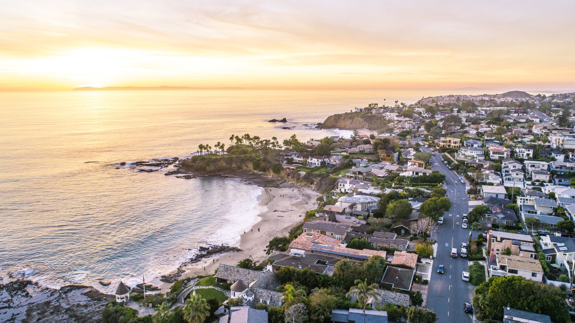 Aerial view of Laguna Beach, California at sunset, showcasing the scenic coastline, sandy beaches, turquoise ocean, and a residential area nestled along the cliffs in Orange Coun