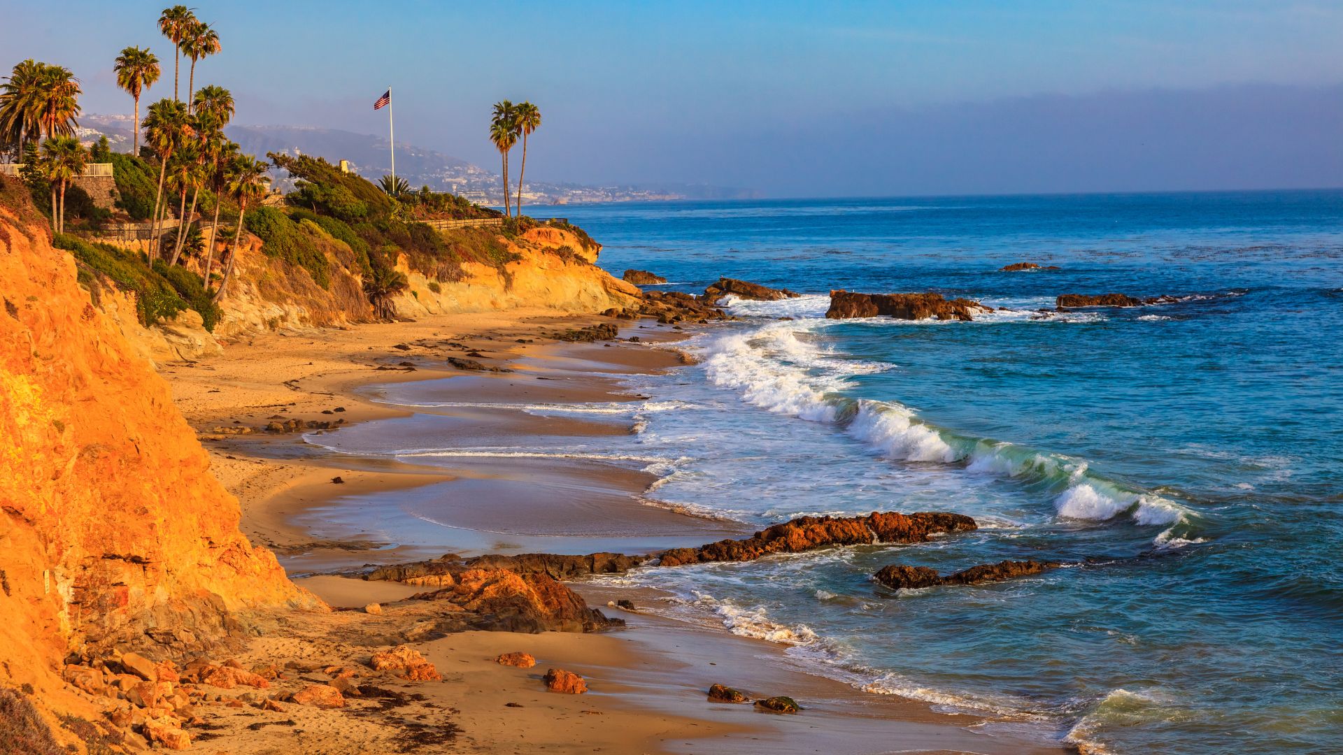 A scenic view of Laguna Beach, California, showcasing a sandy shoreline with waves crashing against rocky outcrops. The beach is flanked by golden cliffs on the left and palm trees on a verdant hillside to the right, under a clear blue sky with a hint of haze over the distant ocean.