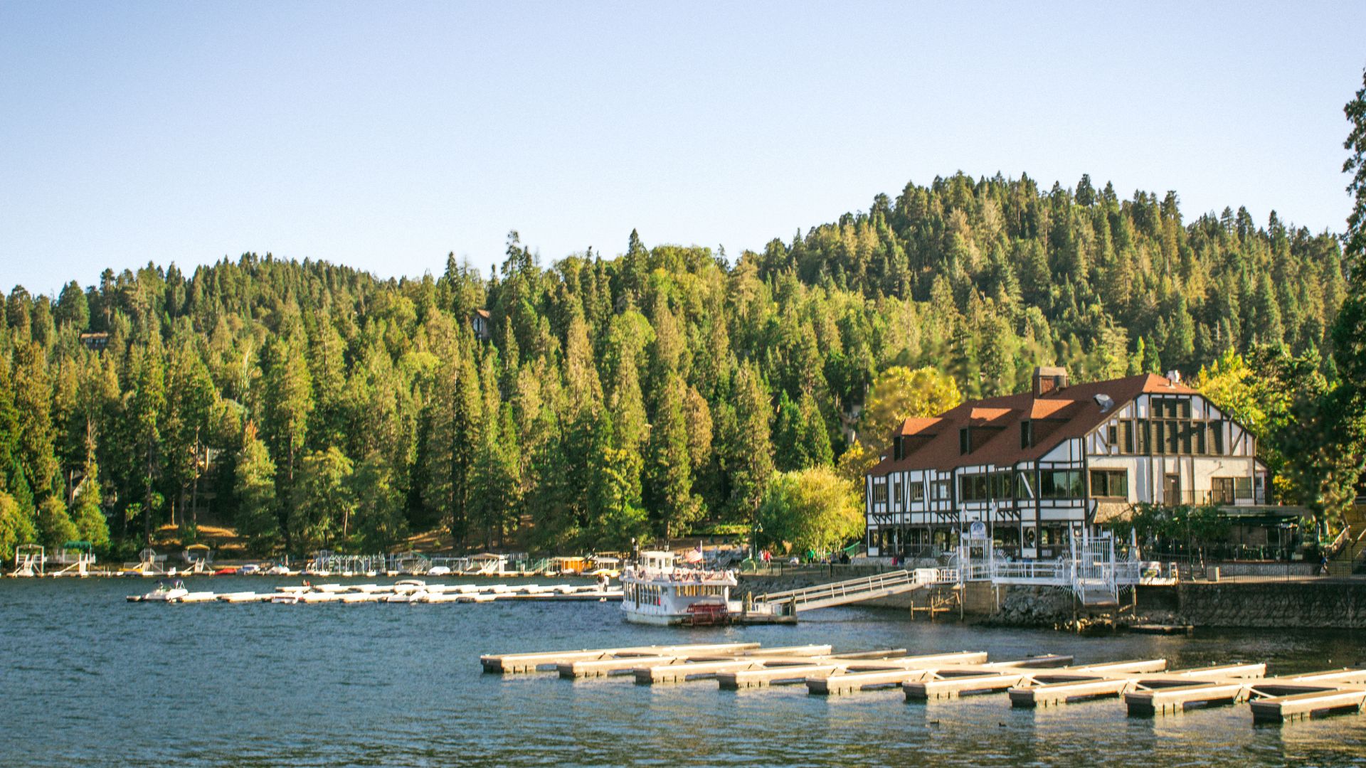 Lake Arrowhead Village in Southern California. View of a serene lake surrounded by a lush forest, with a charming village featuring a distinctive building with a red roof and docks with boats.