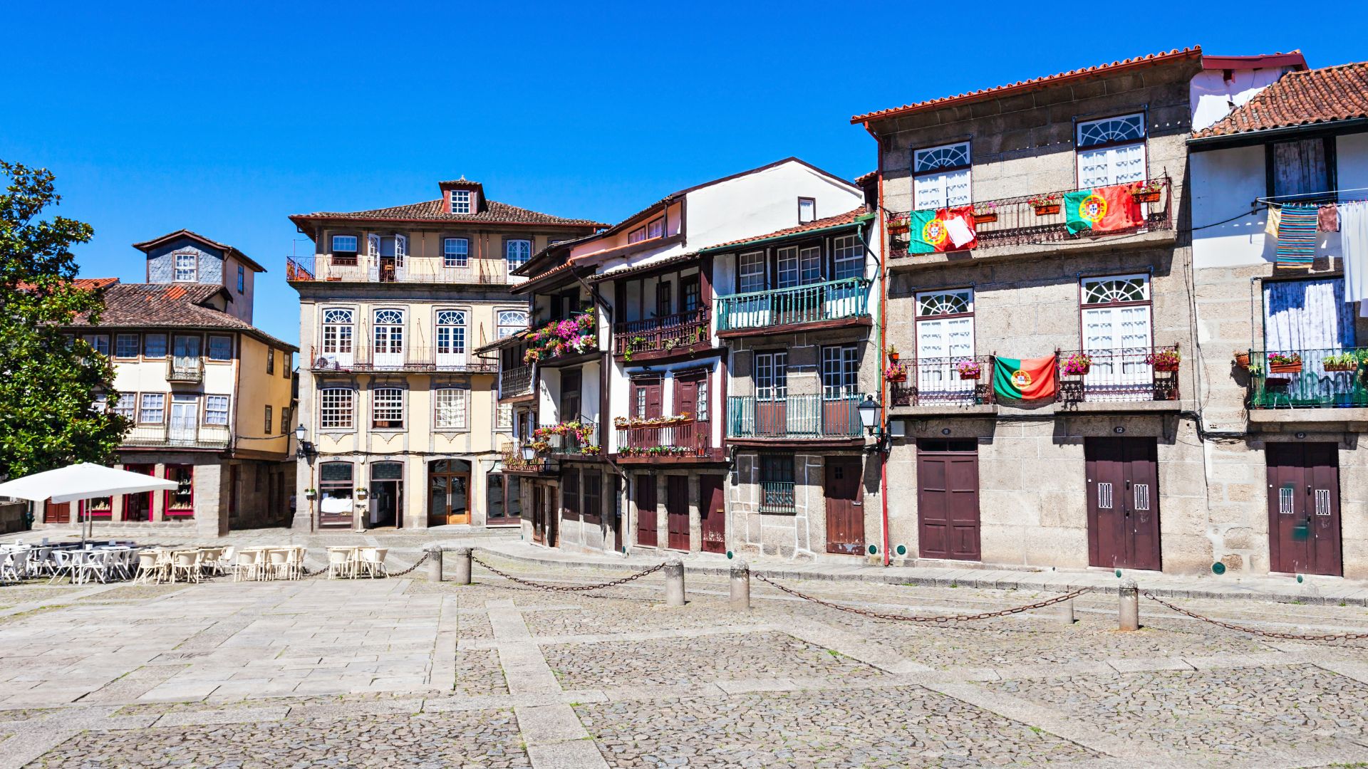 A cobblestone square in Guimarães, Portugal, lined with historic multi-story buildings and a prominent church tower in the background, under a clear blue sky.
