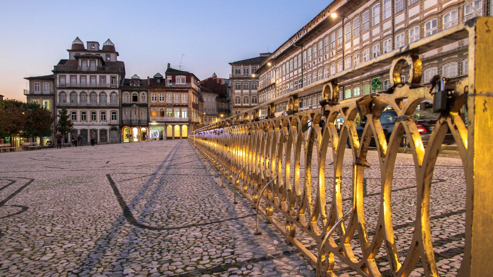 A wide-angle view of Largo do Toural in Guimarães, Portugal, at dusk, showing the illuminated buildings surrounding the square and a golden railing along the foreground.