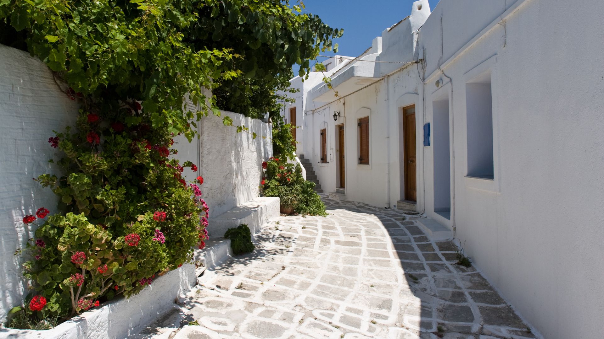 A narrow cobblestone street in Lefkes, Paros, with white buildings and greenery.