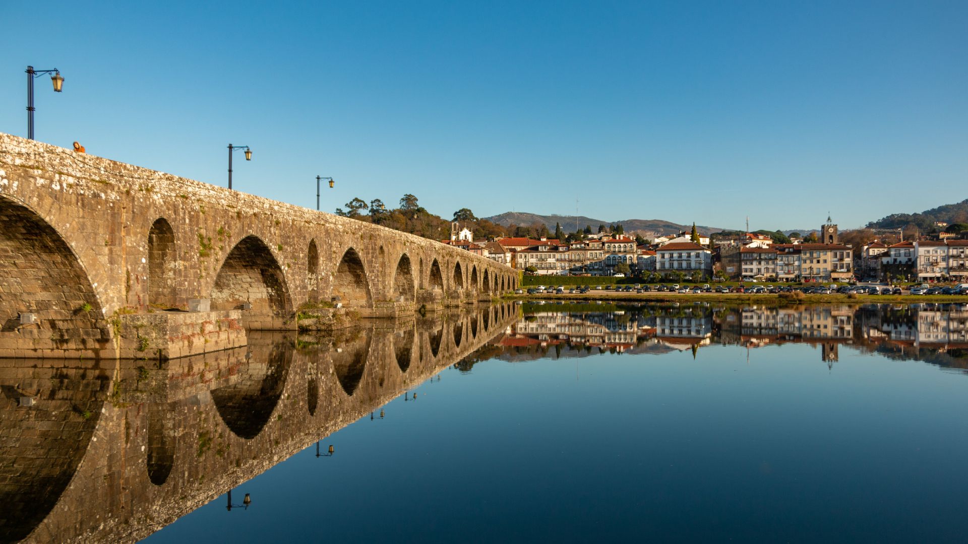 A wide-angle shot of the historic Roman and Gothic bridge in Ponte de Lima, Portugal, reflected in the calm waters of the Lima River, with the town visible on the far bank under a clear blue sky.