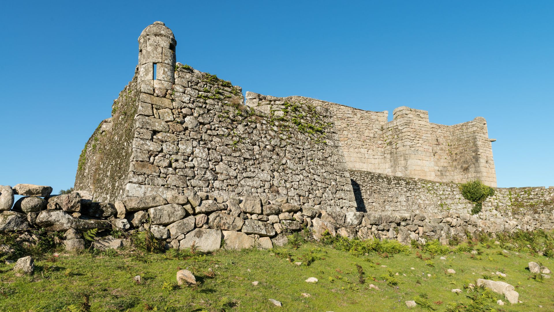 A medieval stone castle with a prominent watchtower stands on a grassy hill under a clear blue sky.