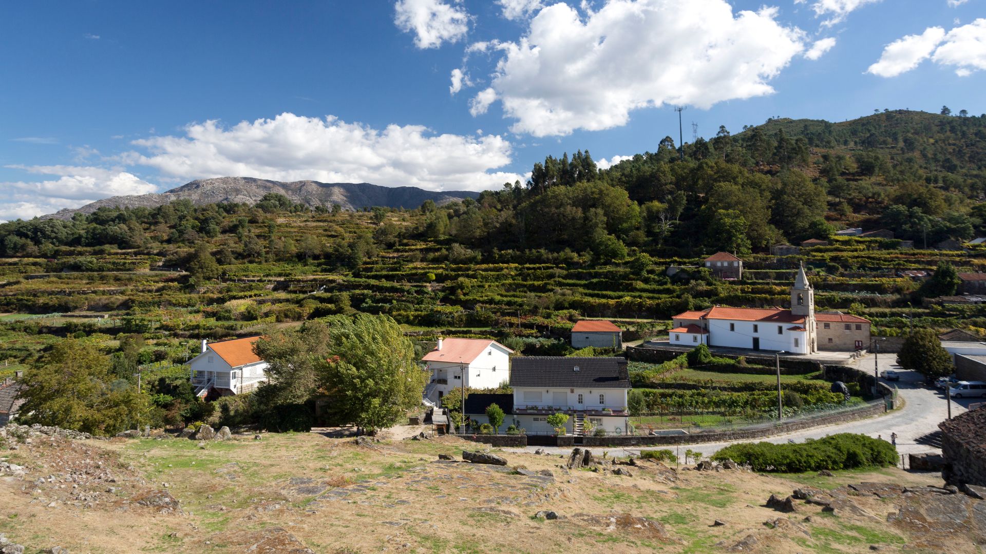 A scenic view of a traditional Portuguese village with white houses and a church surrounded by terraced green fields and a mountainous landscape under a blue sky with white clouds.