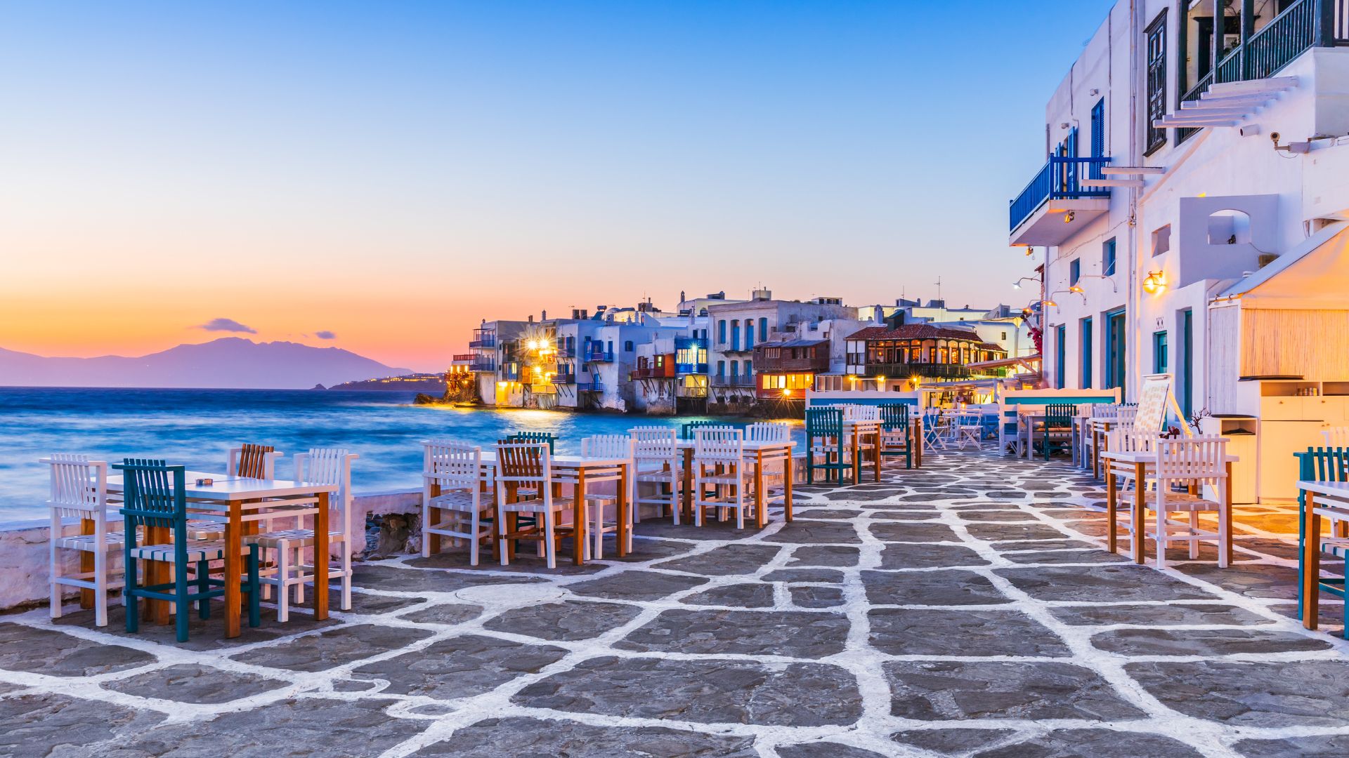 A waterfront cafe scene in Mykonos at sunset, with colorful houses and tables.