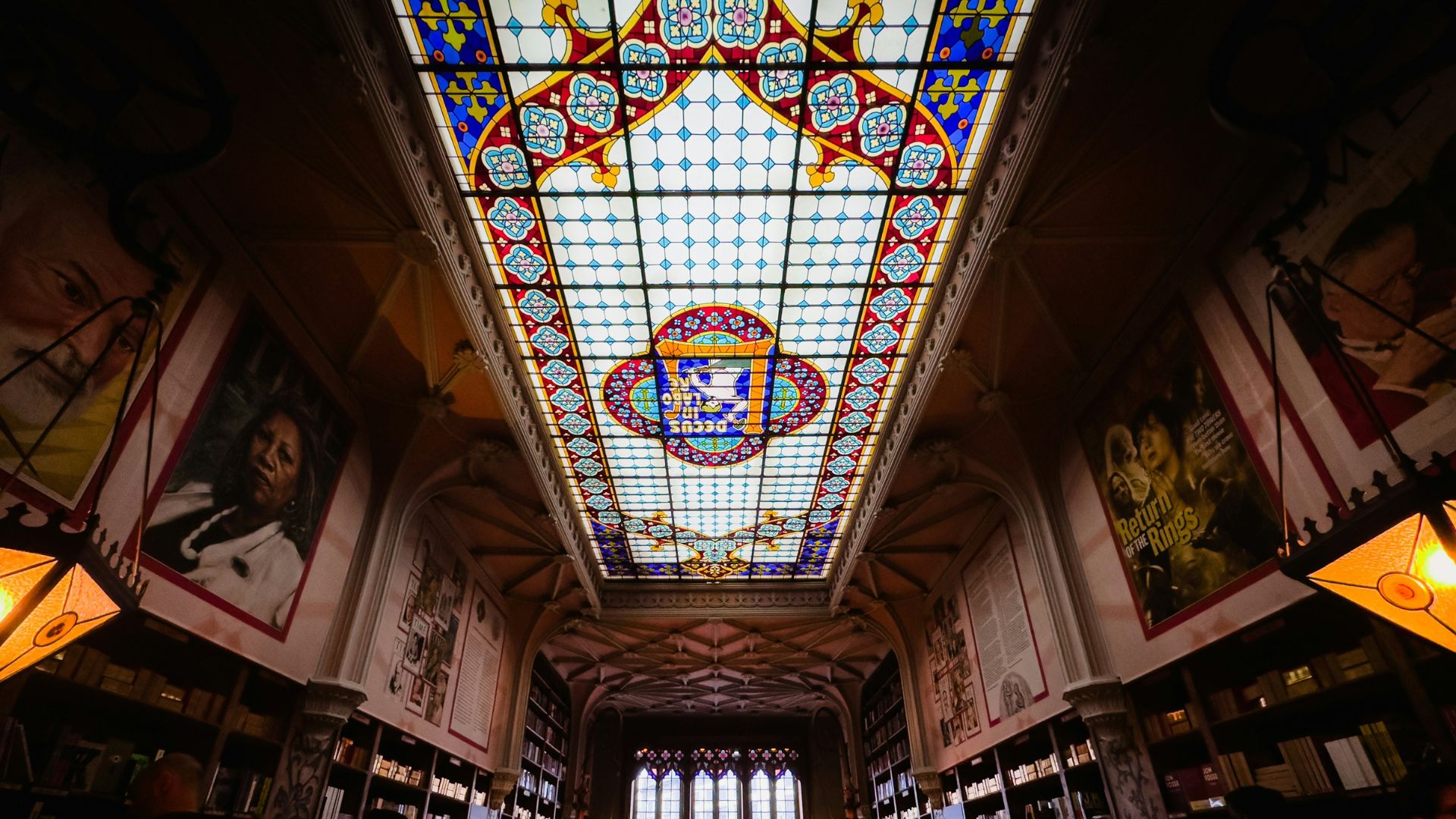 A low-angle shot of the interior of Livraria Lello featuring a large stained-glass ceiling with ornate patterns and bookshelves lining the walls.
