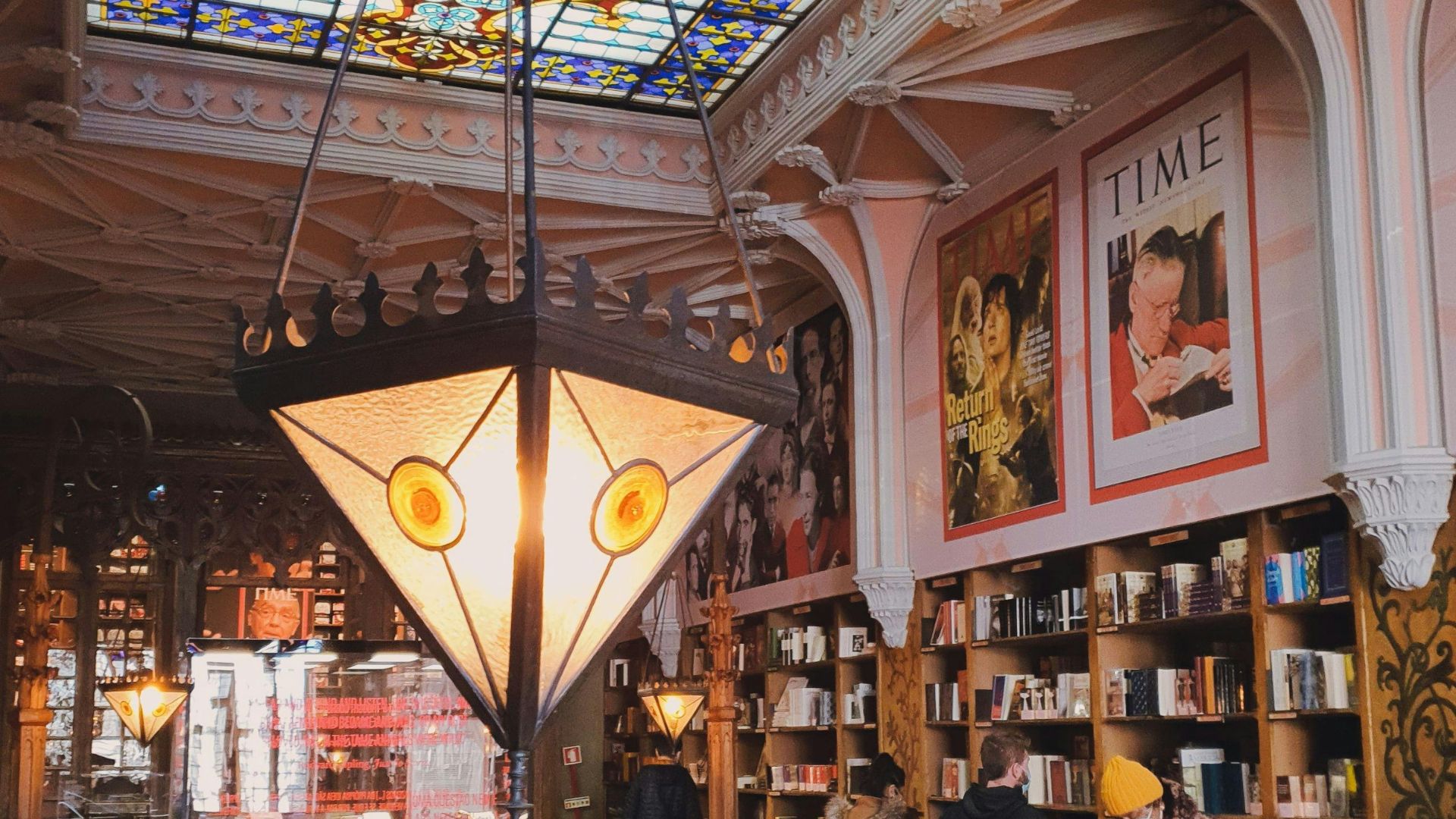 A photo of the interior of Livraria Lello, featuring a large, ornate light fixture hanging from a high ceiling with stained glass, and bookshelves lining the walls.