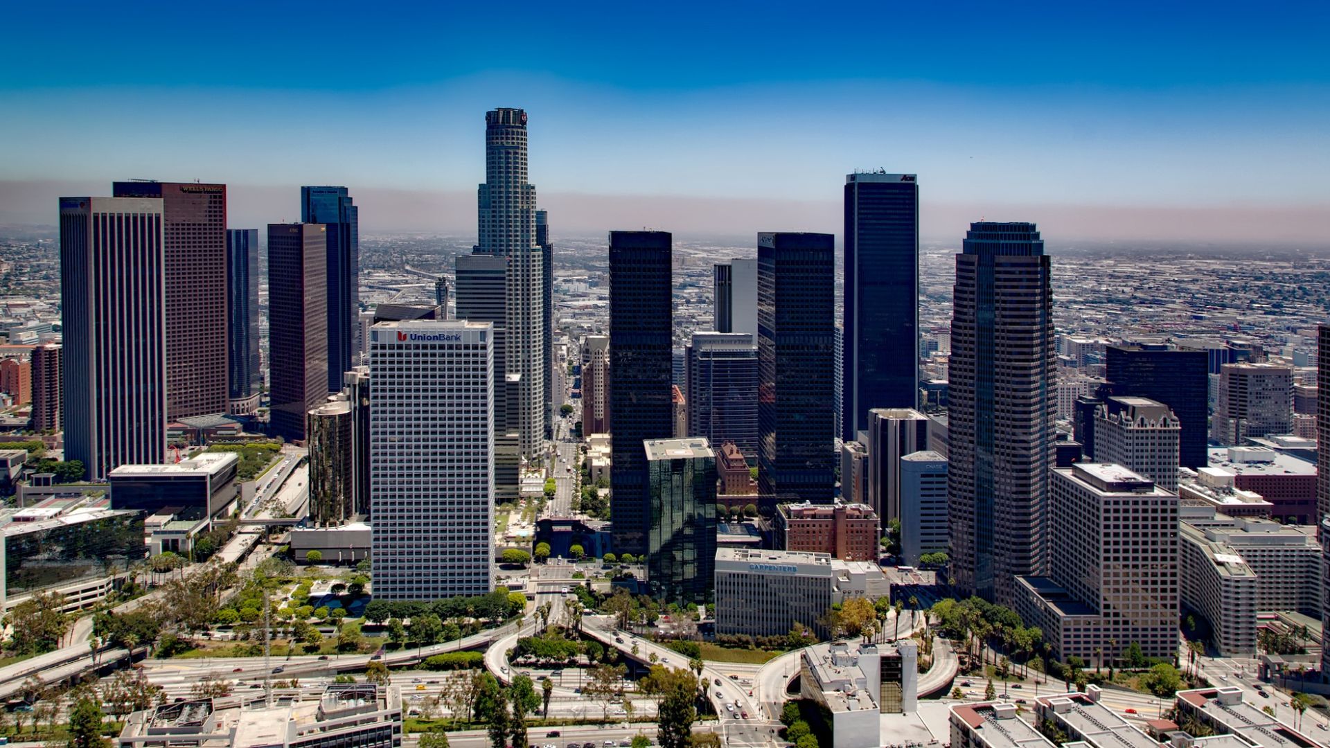 An aerial view of downtown Los Angeles showcasing its towering skyscrapers and bustling freeways under a clear blue sky. The image highlights the city's urban landscape and the iconic U.S. Bank Tower and Union Bank Plaza.