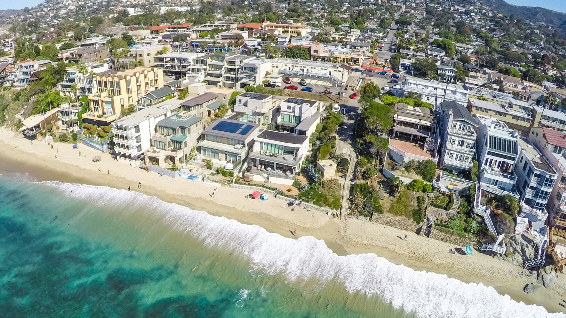 Aerial view of Main Beach in Laguna Beach, California, showcasing the sandy coastline, turquoise ocean, and beachfront properties with lush hills in the background.