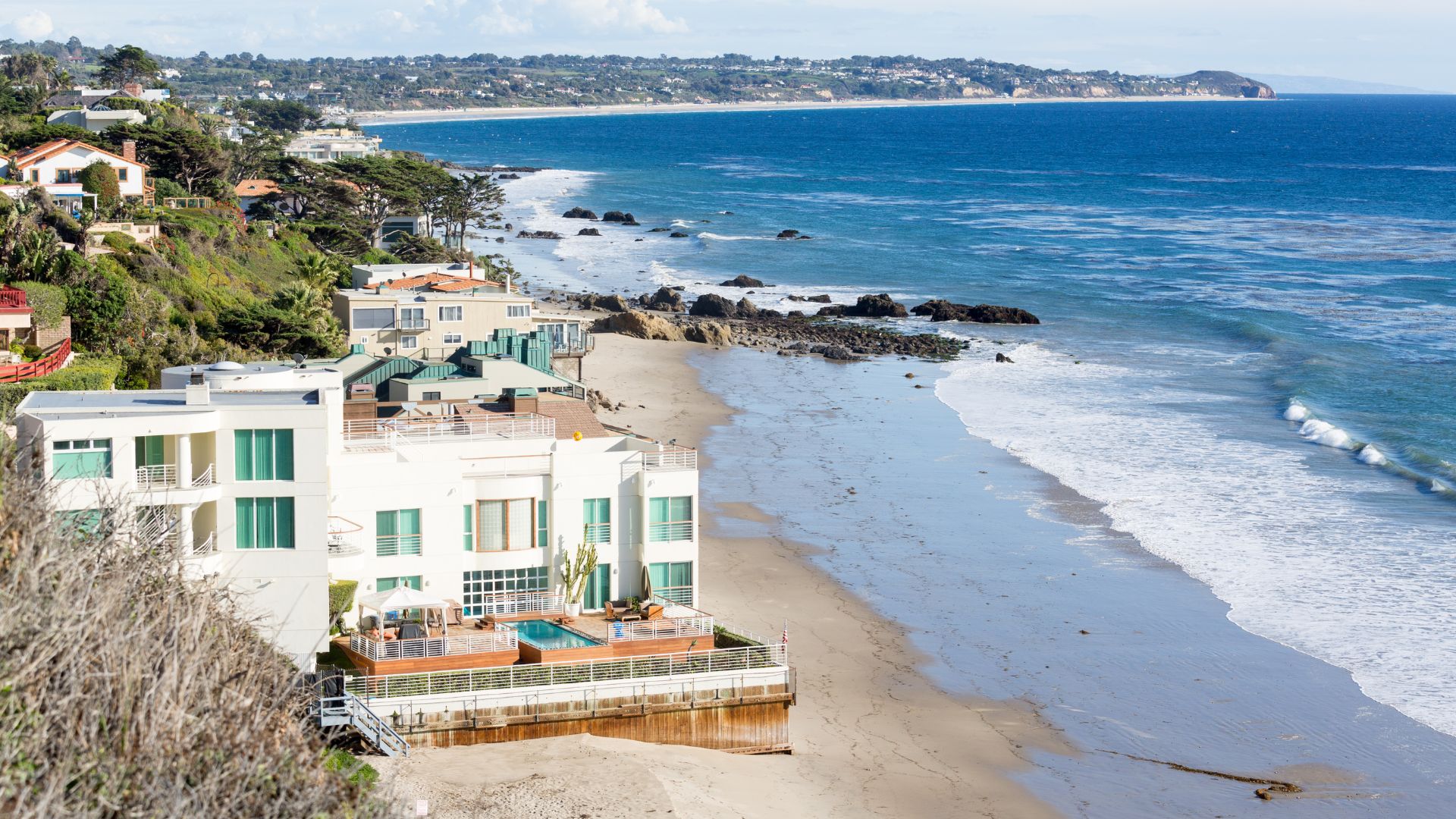 Luxury beach house and coastline in Malibu, California, with the Pacific Ocean and Santa Monica Mountains in the background.