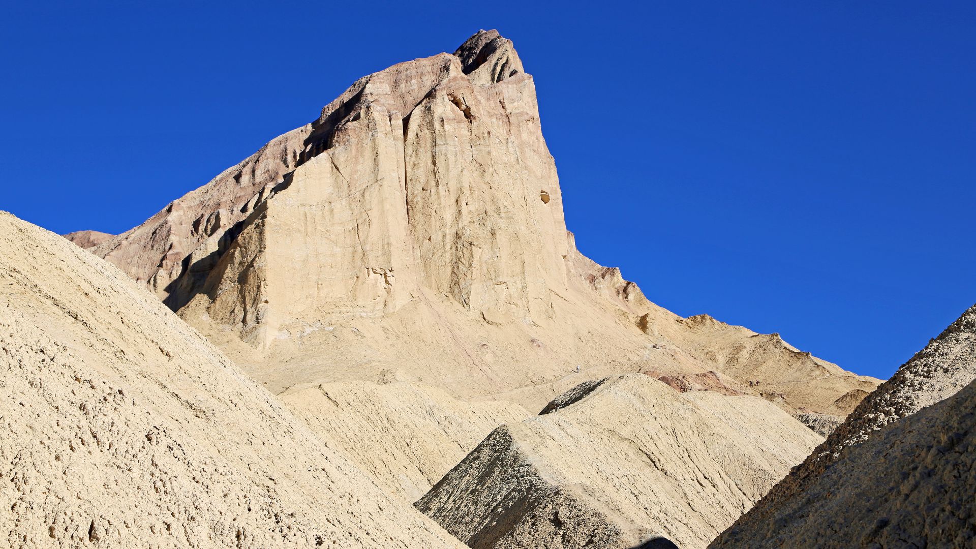 A striking desert landscape under a clear blue sky, featuring Manly Beacon, a tall, light-colored, jagged peak composed of eroded badlands formations, rising sharply from a base of smaller, similarly colored hills in Death Valley National Park.