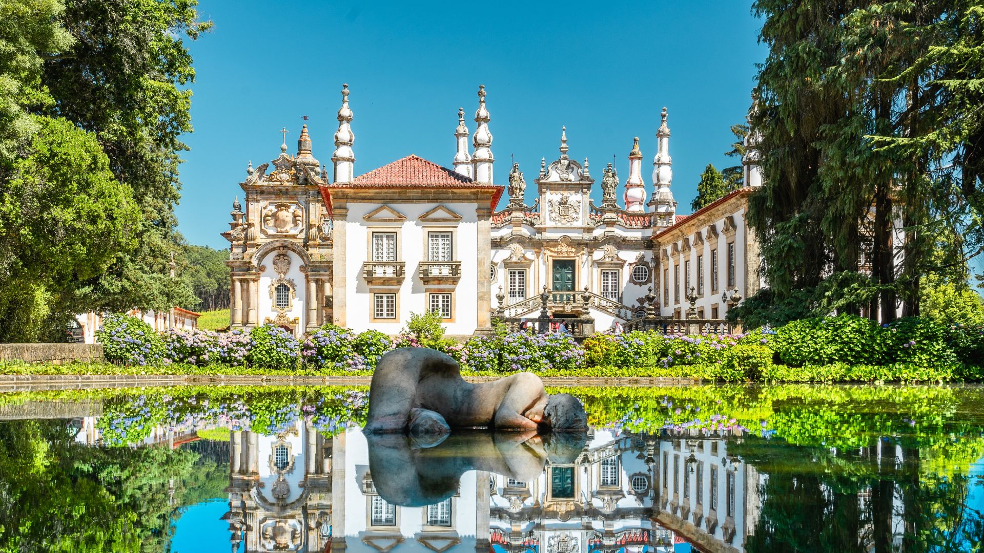 An ornate 18th-century Baroque palace, Mateus Palace, is reflected in a still pond with a modern sculpture of a reclining figure in the foreground, surrounded by lush green gardens and trees under a clear blue sky. 