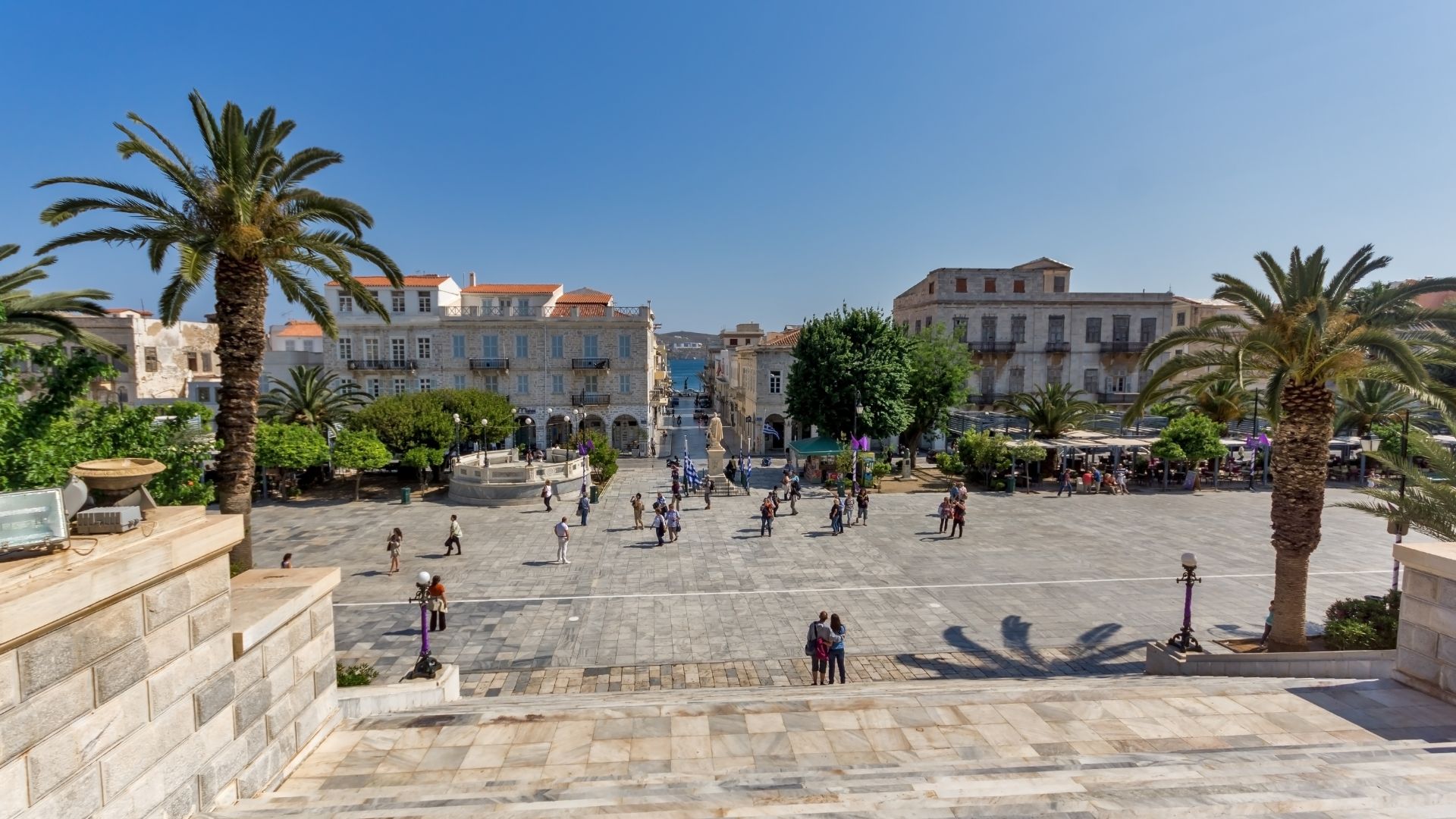 Miaouli Square in Ermoupolis, Syros, Greece, featuring neoclassical buildings, palm trees, and a central square.