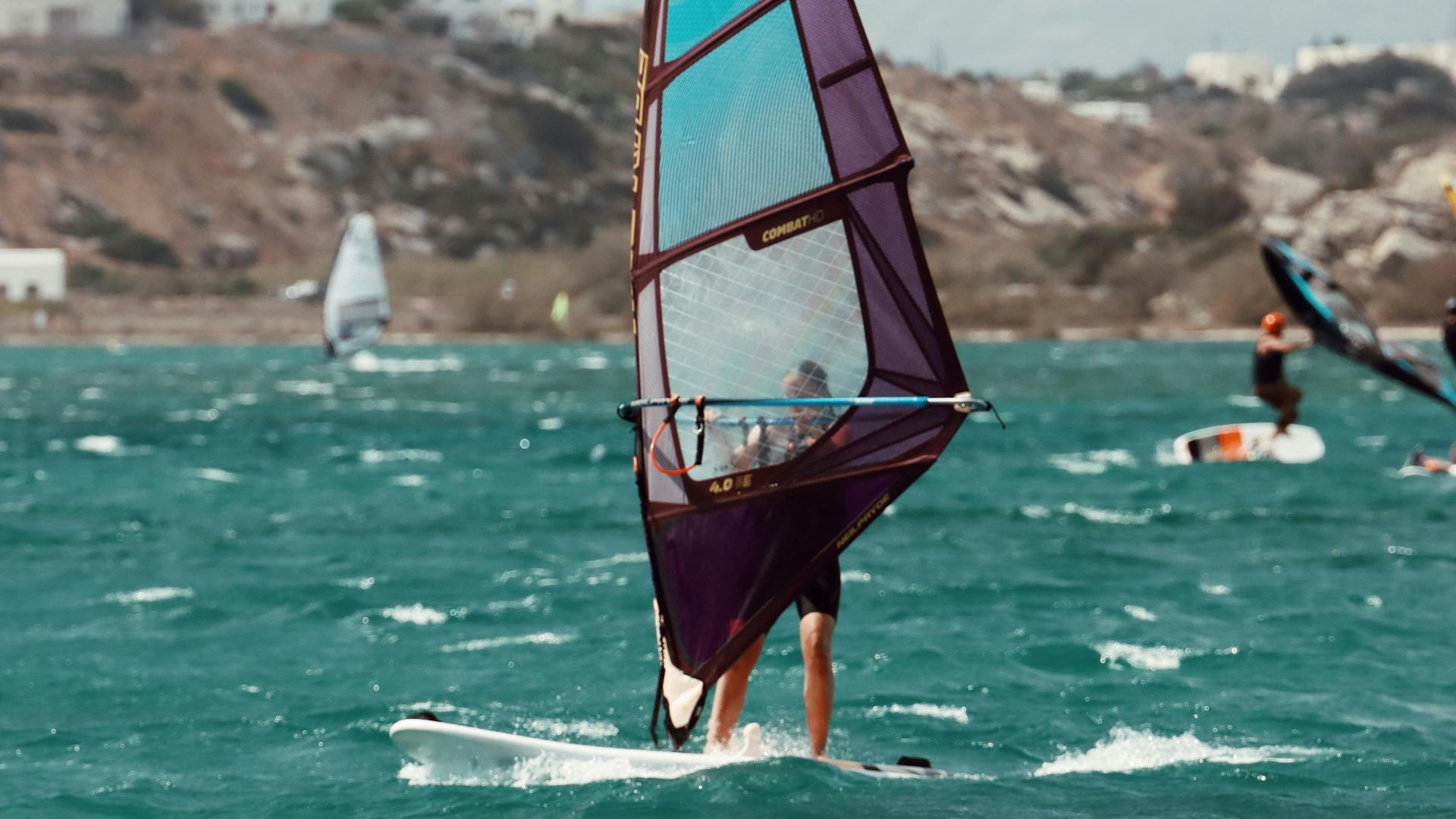 A person windsurfing on a body of water with a blue and purple sail, other windsurfers are in the distance.